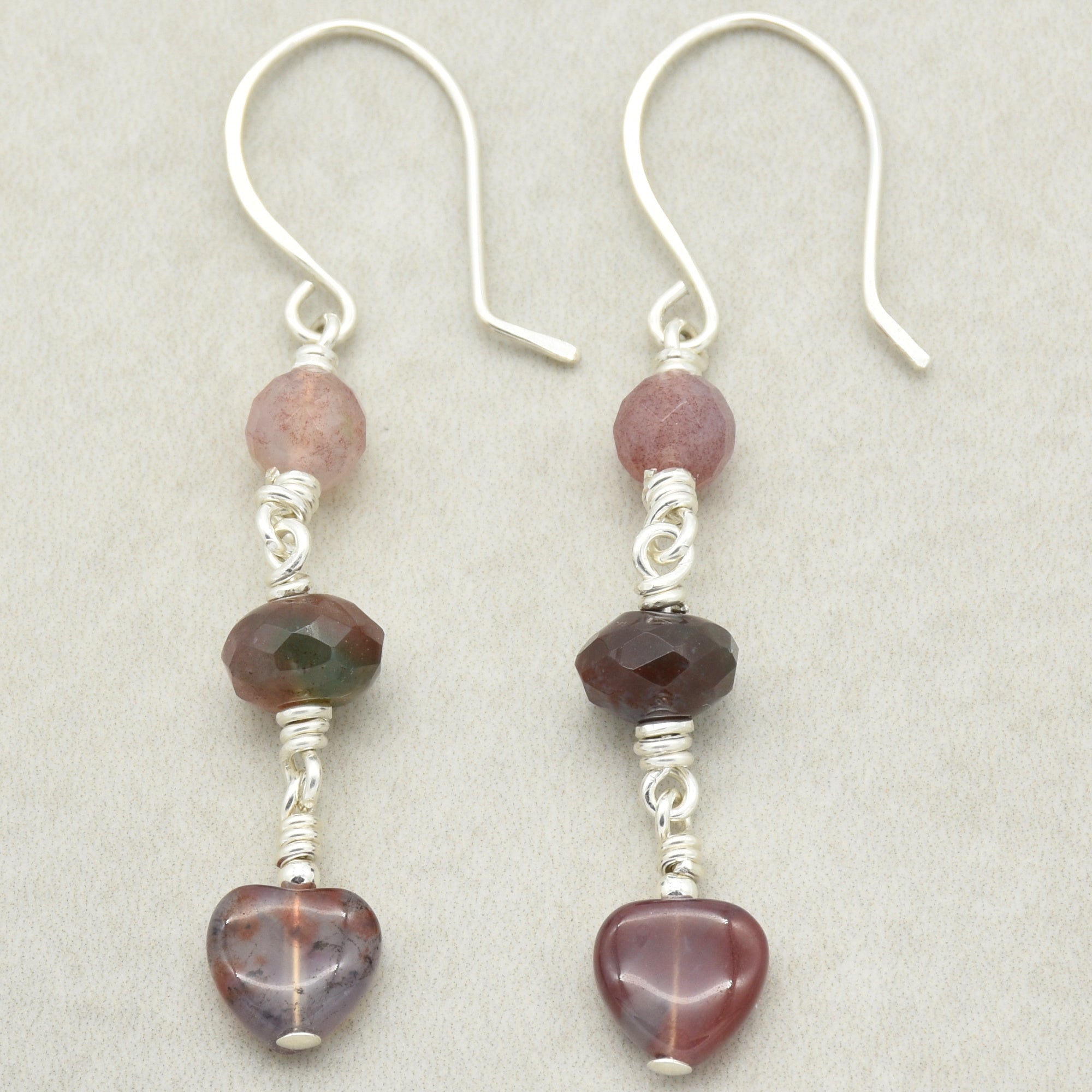 The image shows a pair of earrings. They have hooks for the ears and feature three beads each, including a heart-shaped bead at the bottom.