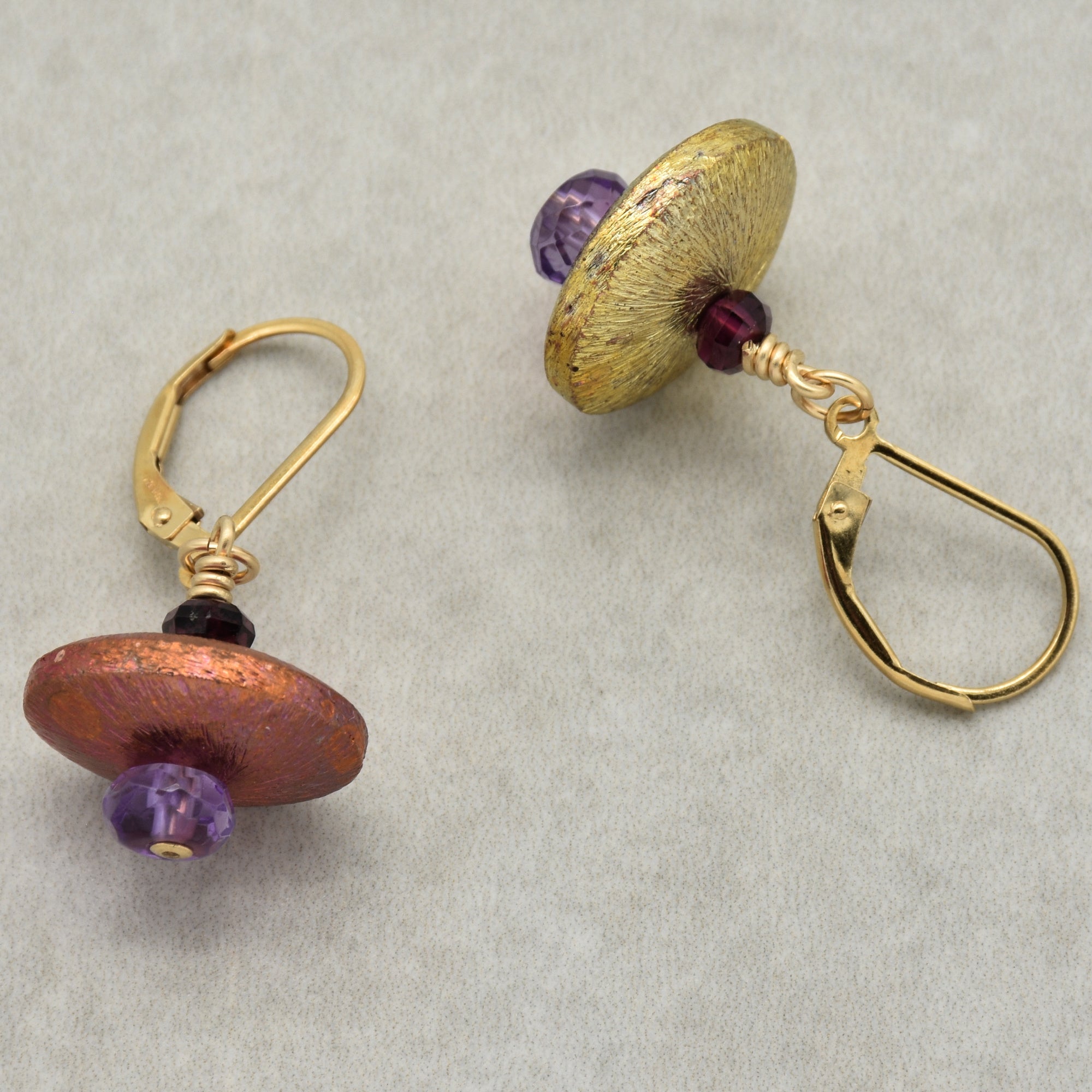 The image shows a pair of earrings. They have lever-back hooks and decorative elements with circular, disk-like beads in gold and copper tones, accented with purple beads.