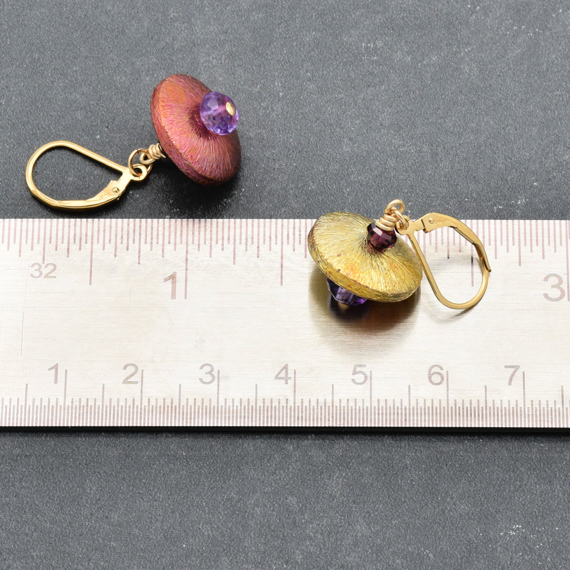 The image shows a pair of earrings. They have a circular design with a central purple bead and a metallic petal-like structure around the bead. The earrings are placed next to a ruler for size reference.