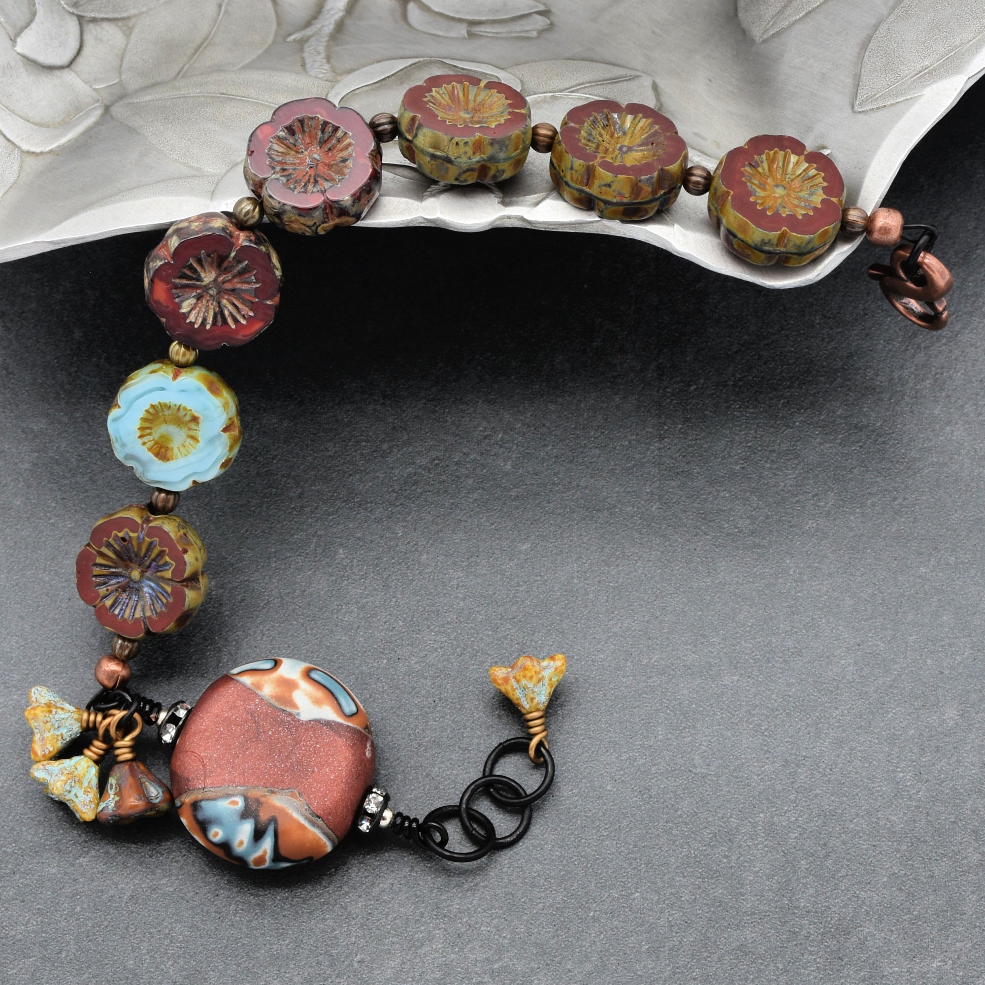 This image shows a bracelet. The jewelry piece is a circular or semi-circular form, which is characteristic of bracelets, and it has a clasp for fastening, visible on the right side. The design includes colorful, flower-shaped beads and a larger decorative bead.