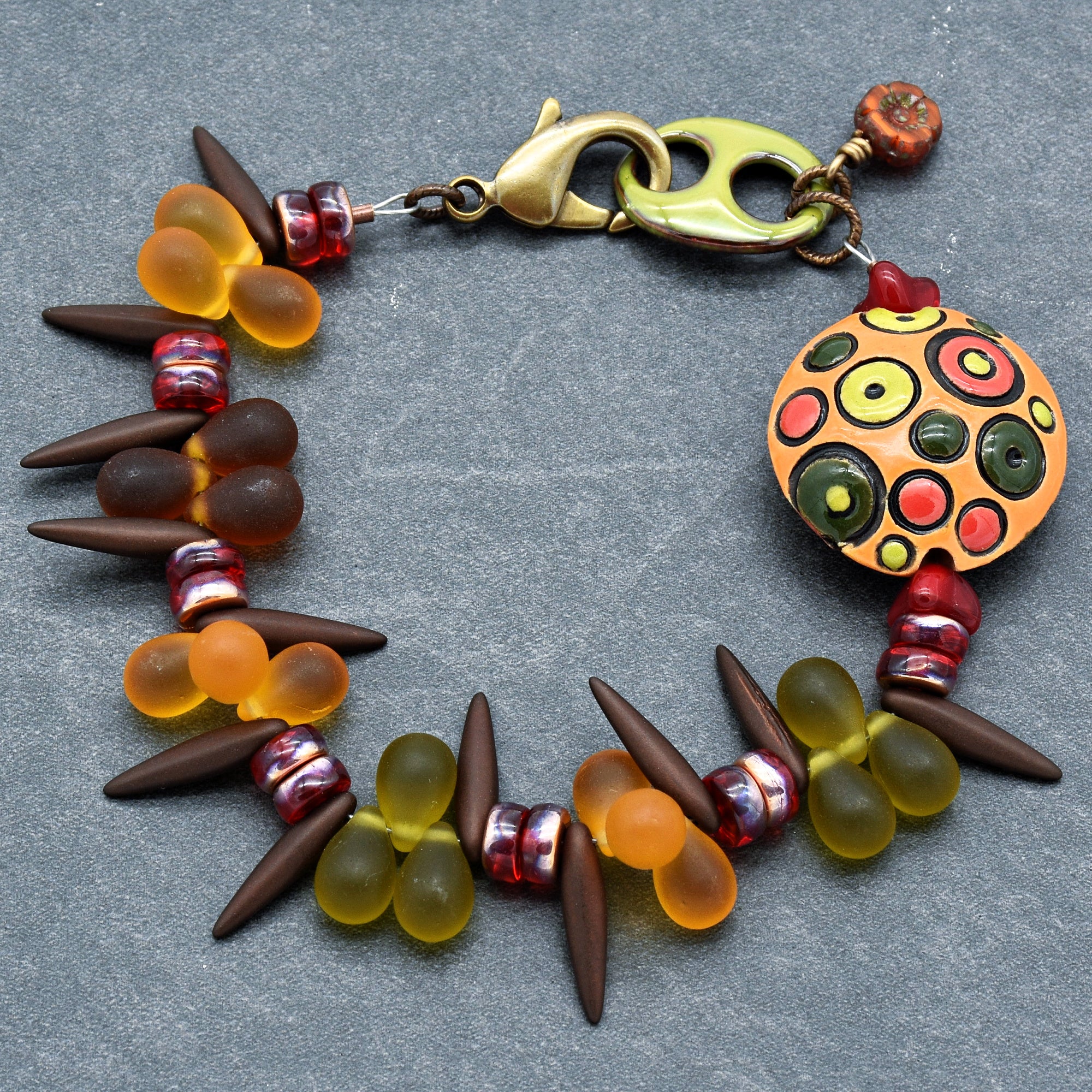 This image shows a bracelet. It features a combination of colorful beads, including orange and green teardrop-shaped beads, elongated brown beads, and smaller round red beads. The bracelet is secured with a metal clasp.