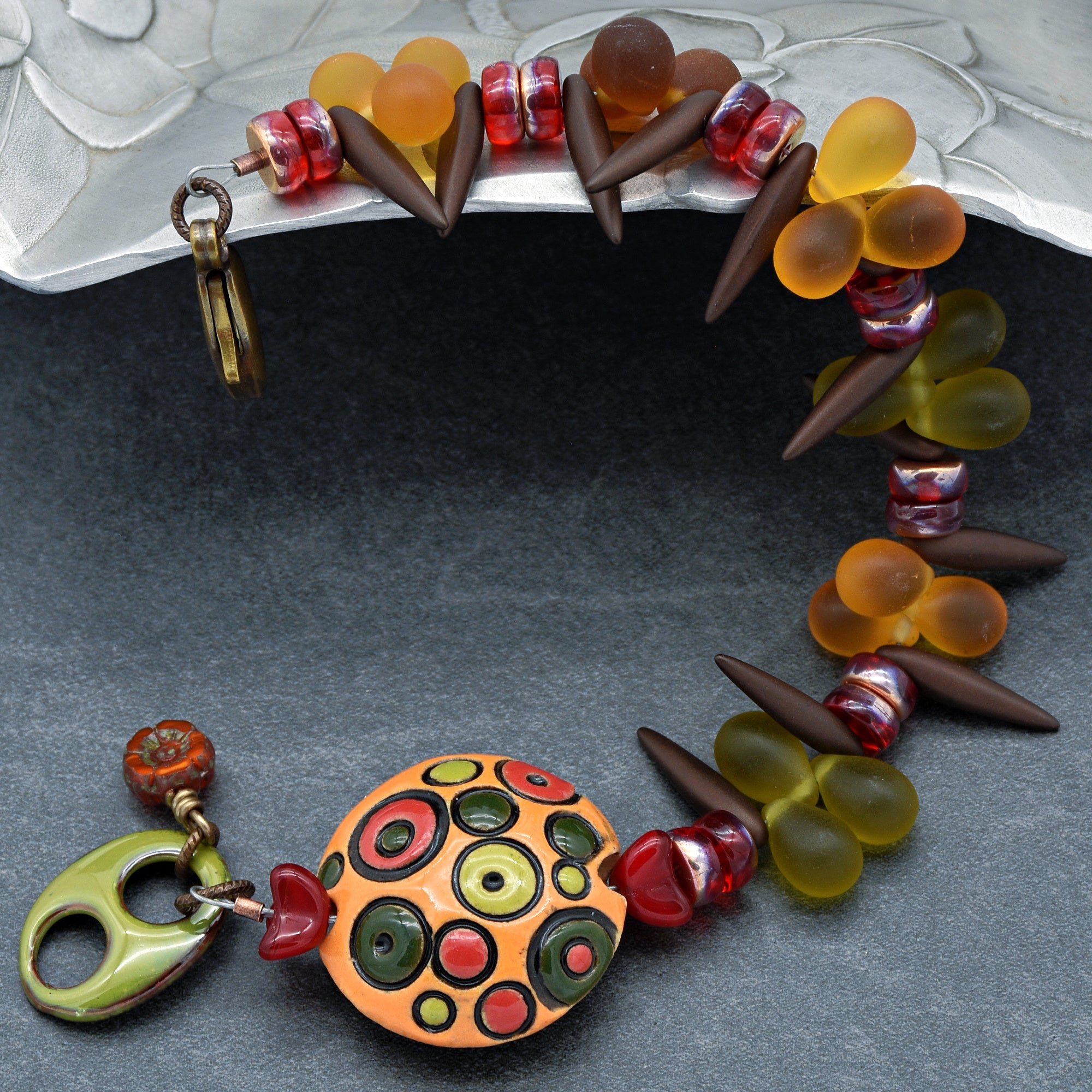 This image shows a bracelet. It features colorful beads in shades of orange, red, and green, with a prominent central bead that has a circular pattern design. The bracelet also includes dark, elongated beads and a clasp for fastening.