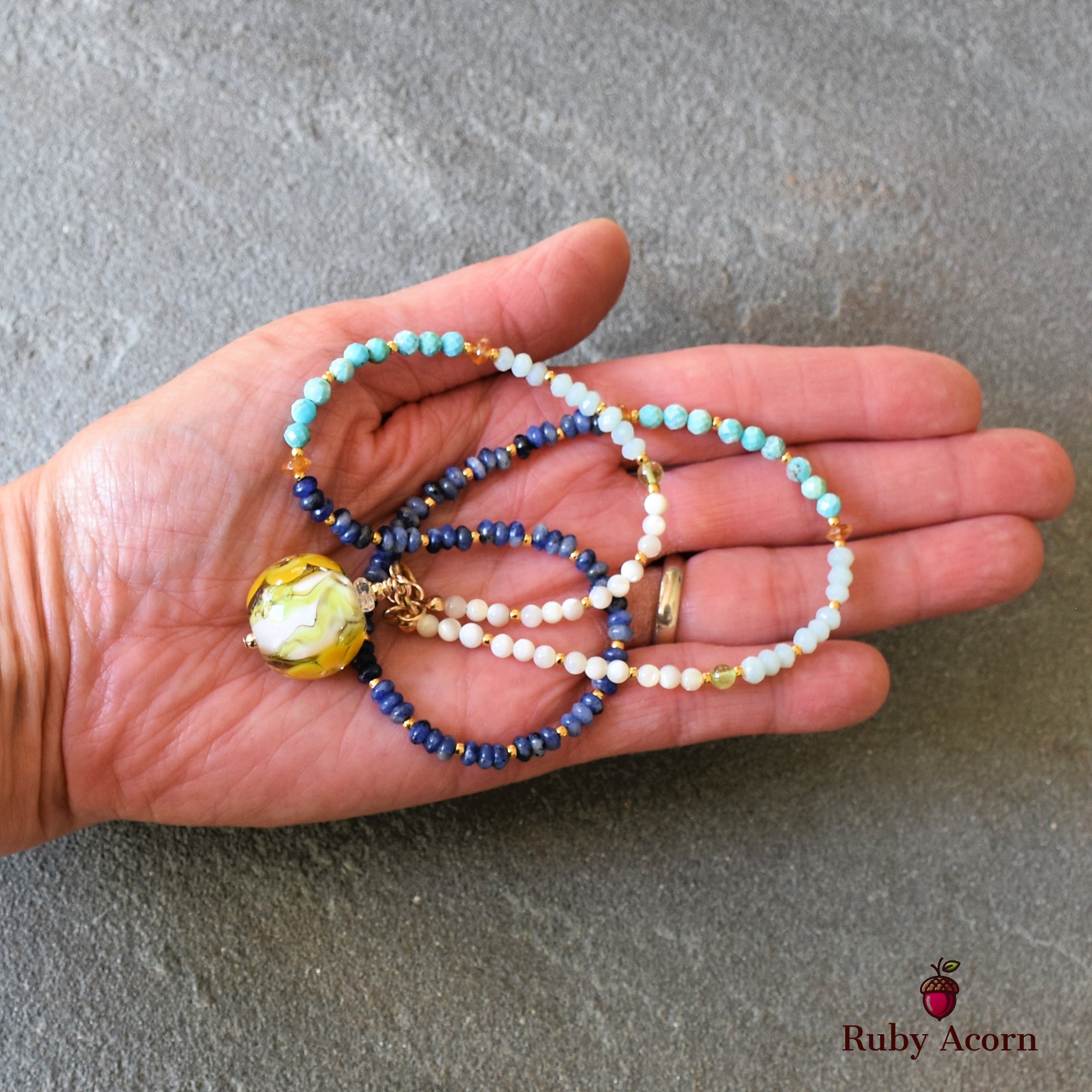 This image shows a necklace. It features strands of beads in various colors (blue, white, turquoise) and has a large round pendant with yellow and green swirls. The item is being held in a person's hand, which helps to show the length and design, consistent with a necklace.
