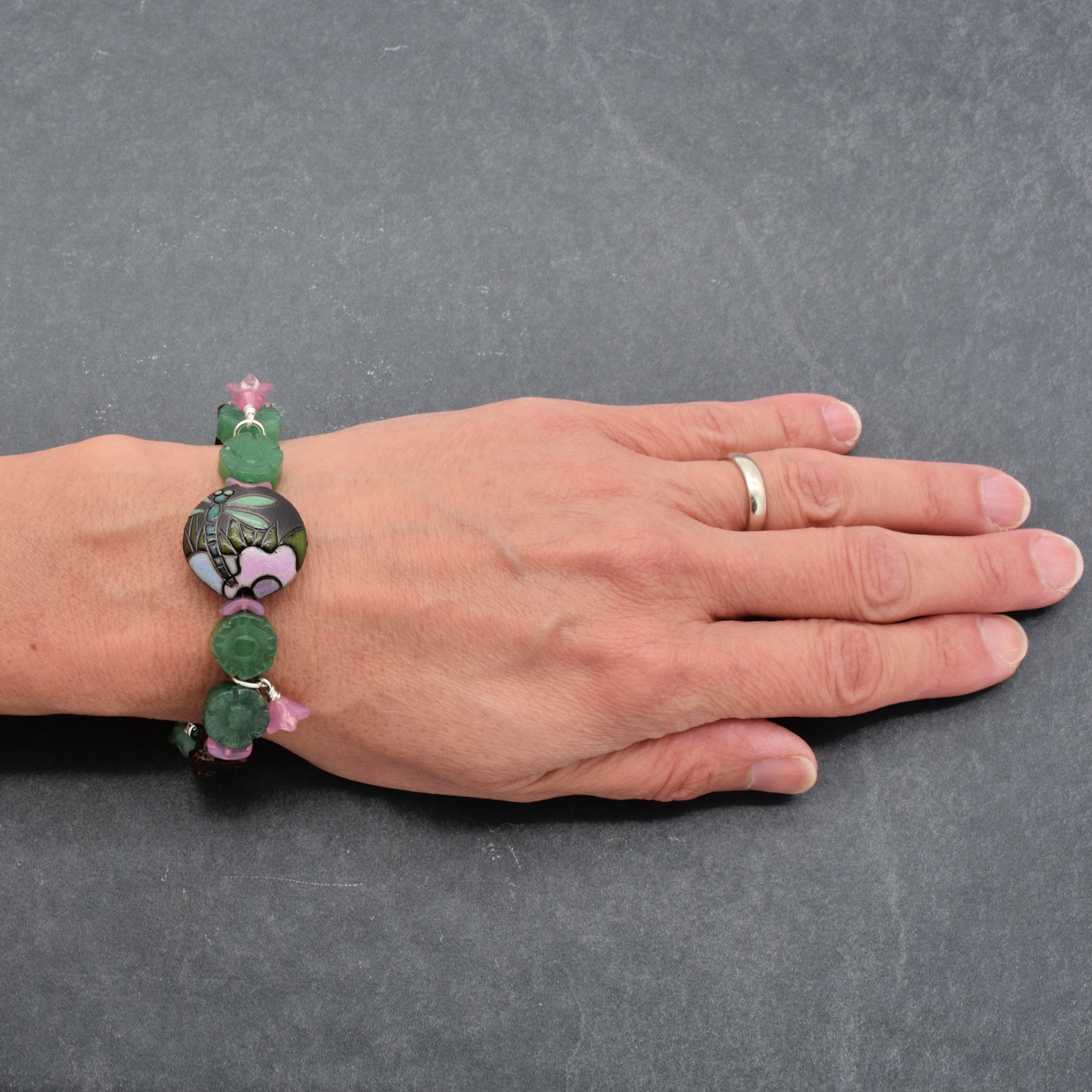 This image shows a bracelet. It is worn around the wrist and features beads with green and pink elements, including a central decorative bead with a floral or natural motif.