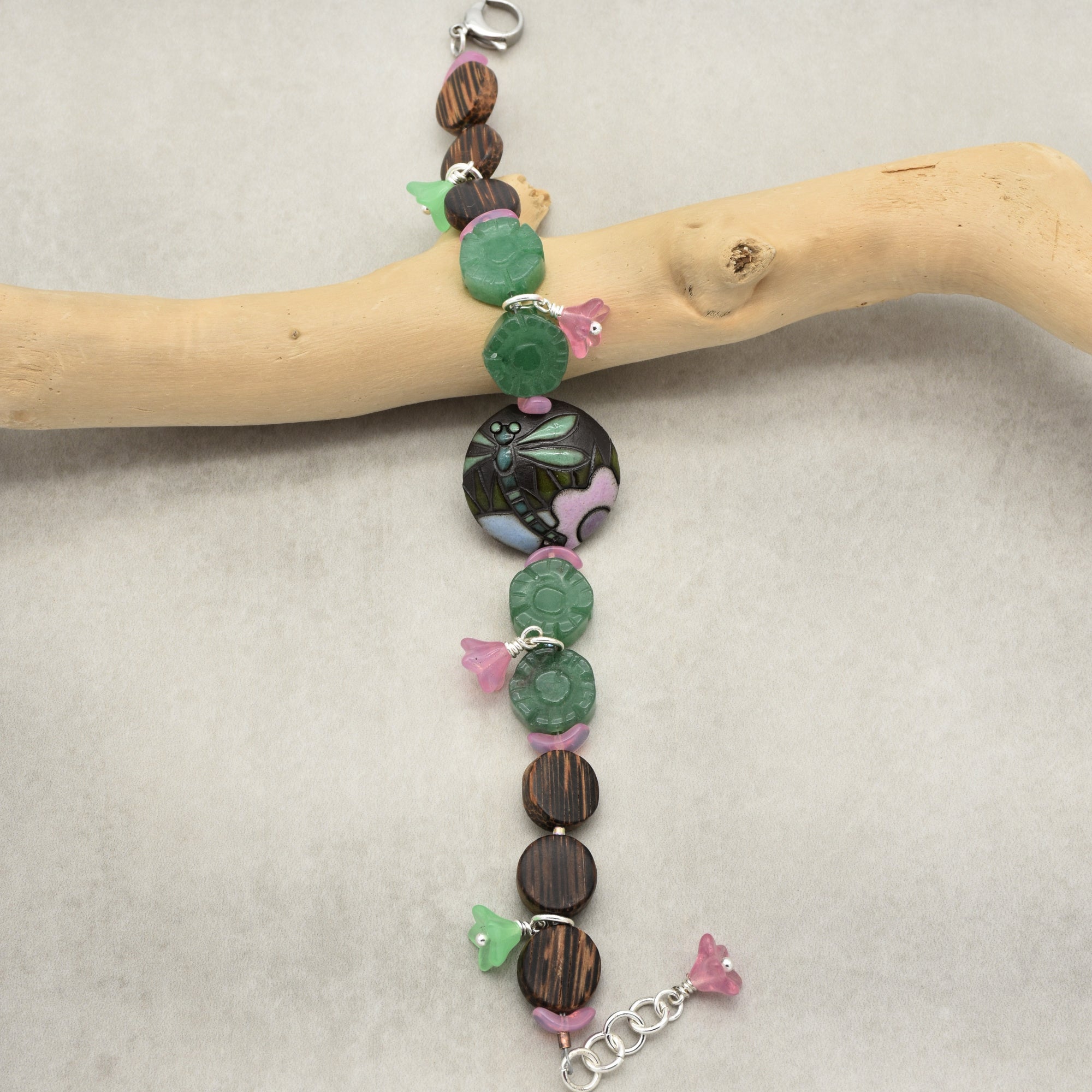 The image shows a bracelet. It has a clasp on one end and an adjustable chain on the other, with a design featuring green and brown beads, pink flower-shaped accents, and a central circular bead with a dragonfly design.