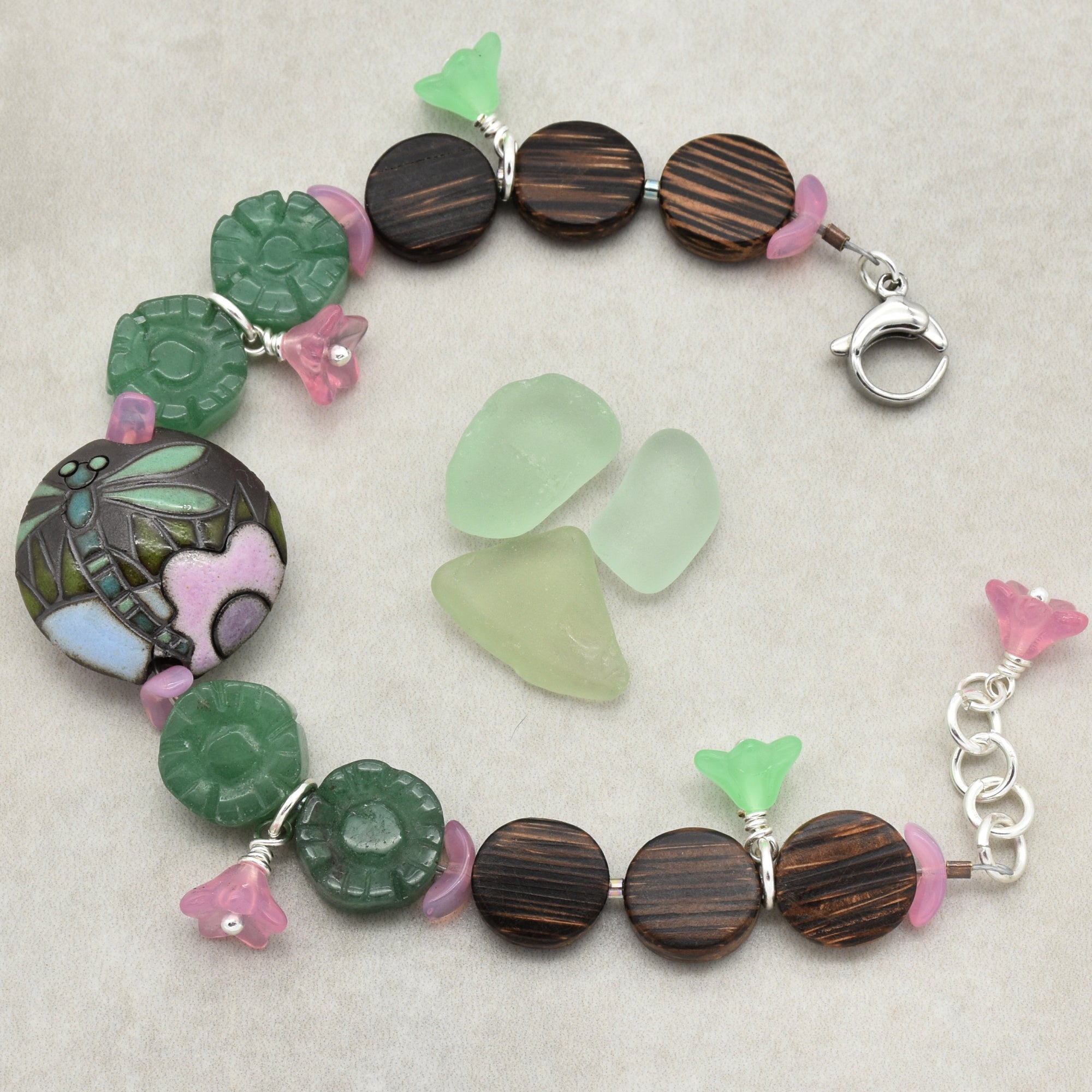 This image shows a bracelet. The jewelry piece forms a circular shape with a clasp, typical for bracelets. It features various beads, including wooden round beads, green carved beads, small pink and green flower-shaped beads, and a larger central bead with a dragonfly and floral design.
