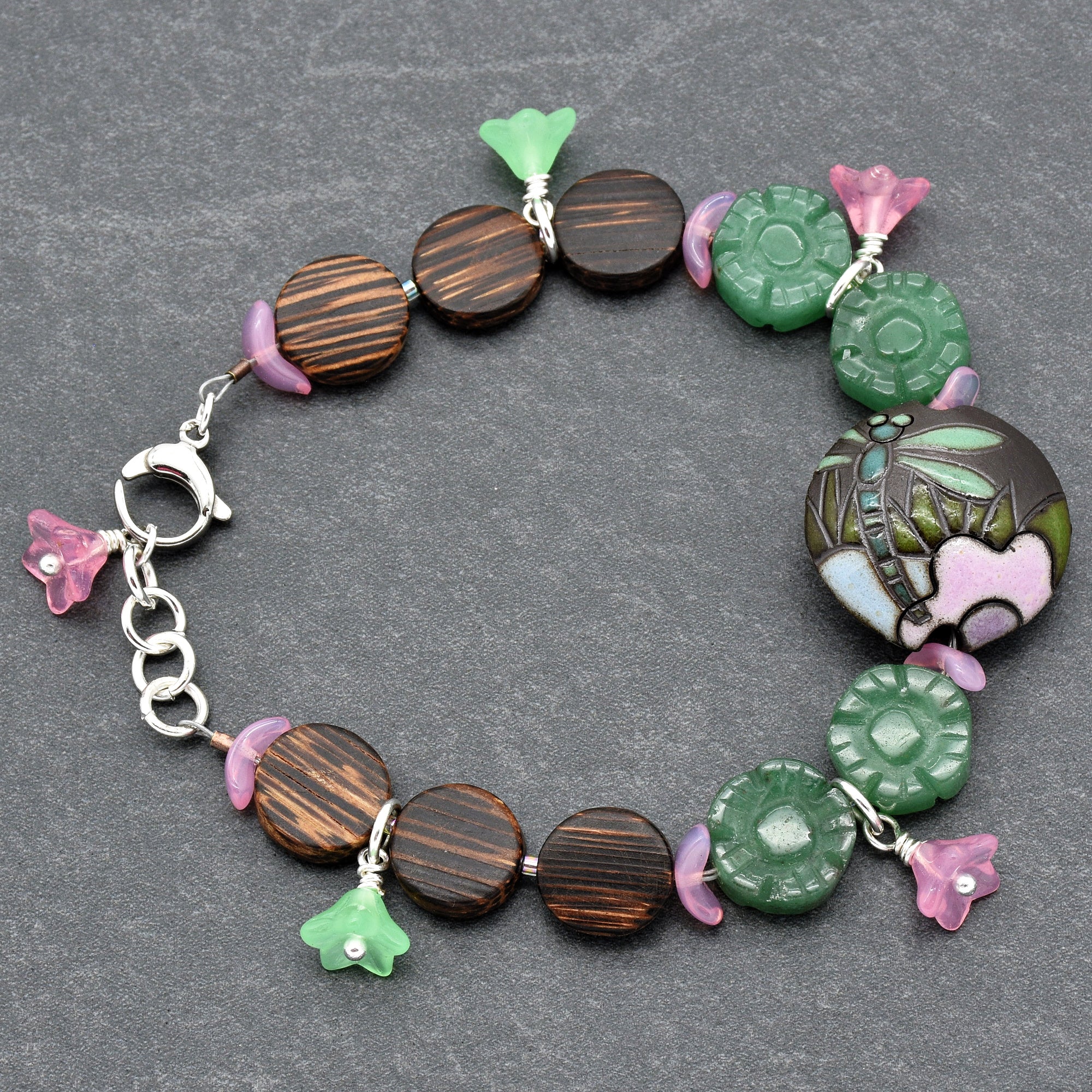 The jewelry in the image is a bracelet. It features a clasp and is designed to fit around the wrist. The bracelet has round beads, some green floral-shaped accents, a large decorative round bead with a dragonfly and flower design, and small pink and green flower charms.
