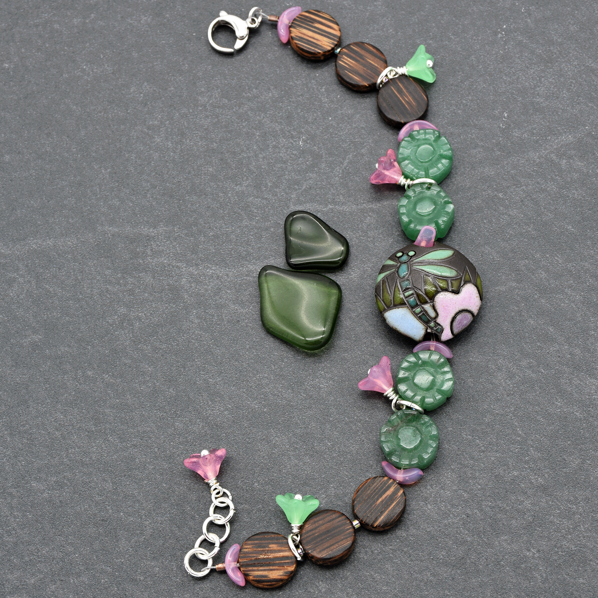 This image shows a bracelet. The bracelet has a clasp and is decorated with beads, including patterned circular beads, green and pink floral-shaped beads, and a central round bead with a dragonfly and flower design. The overall size and the clasp style indicate that it is intended to be worn on the wrist.