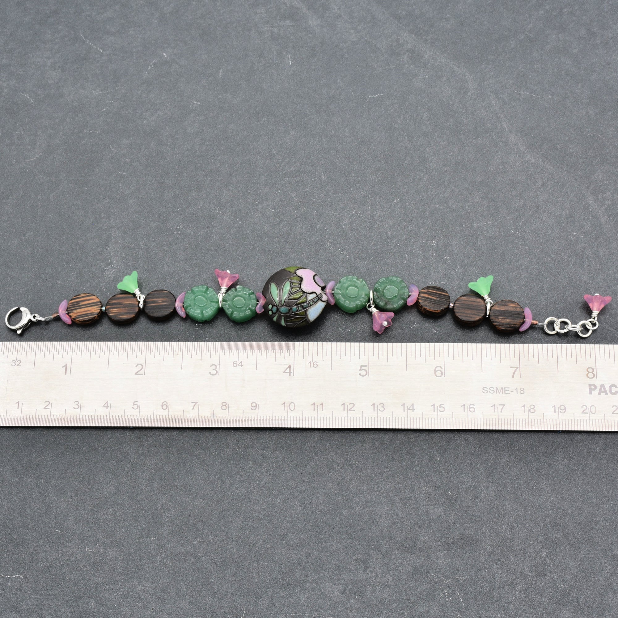 This image shows a bracelet. The jewelry is about 7 inches long as measured against the ruler and has a clasp on one end, which is typical for bracelets. It is composed of green, purple, and brown beads and decorative elements.