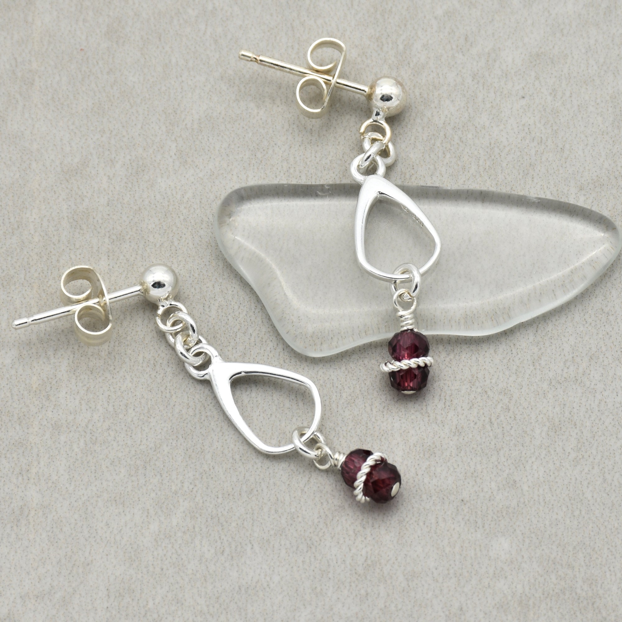 This image shows a pair of earrings. They are stud earrings with a small ball at the top and a dangling design that includes a silver outline shape and a small purple bead wrapped with silver wire.