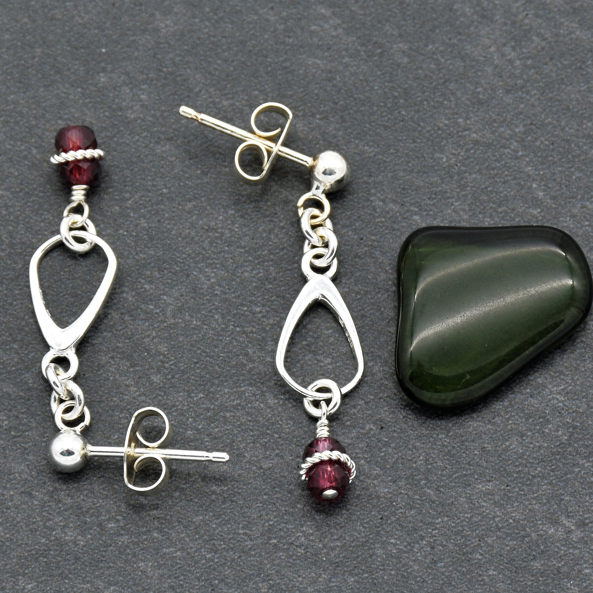 This image shows a pair of earrings. They have a stud post with a butterfly back and a dangling element featuring a small dark red bead and a silver loop.