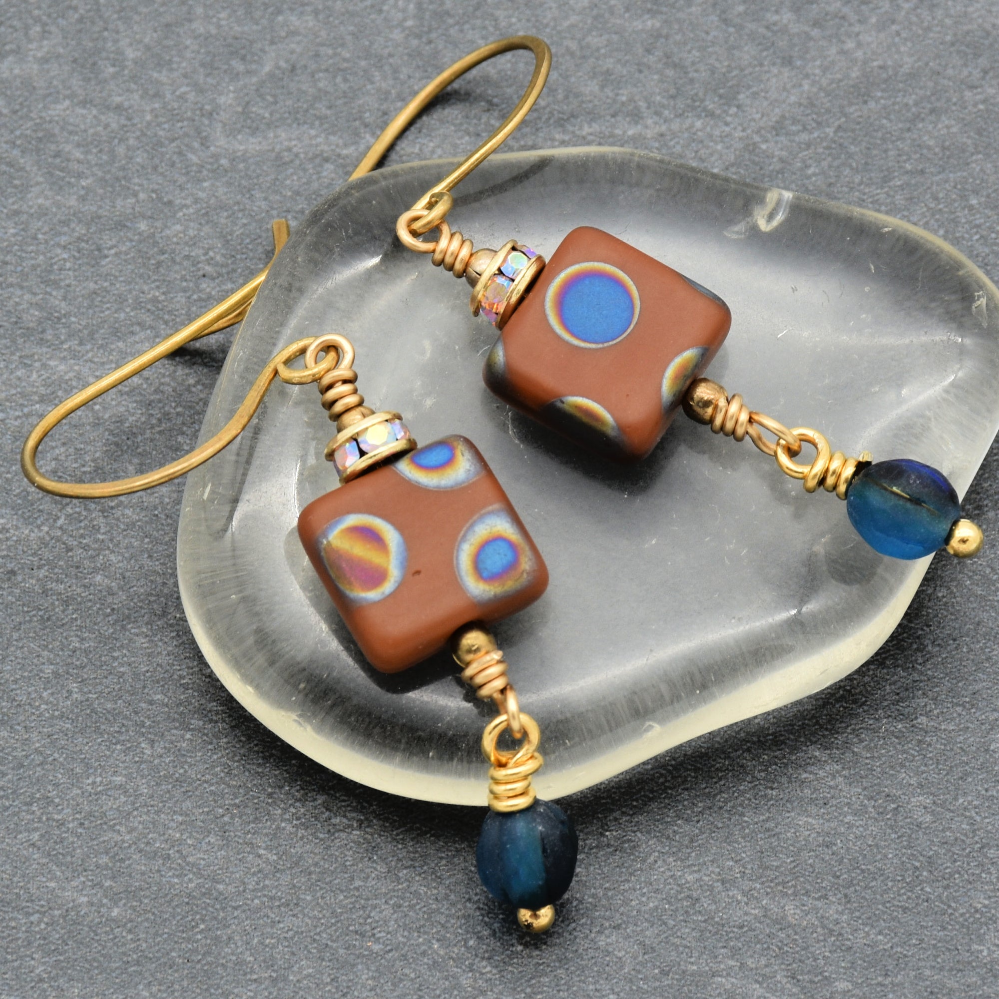 This image shows a pair of earrings. They feature square beads with colorful circular patterns, along with smaller blue beads and gold-toned wire hooks.