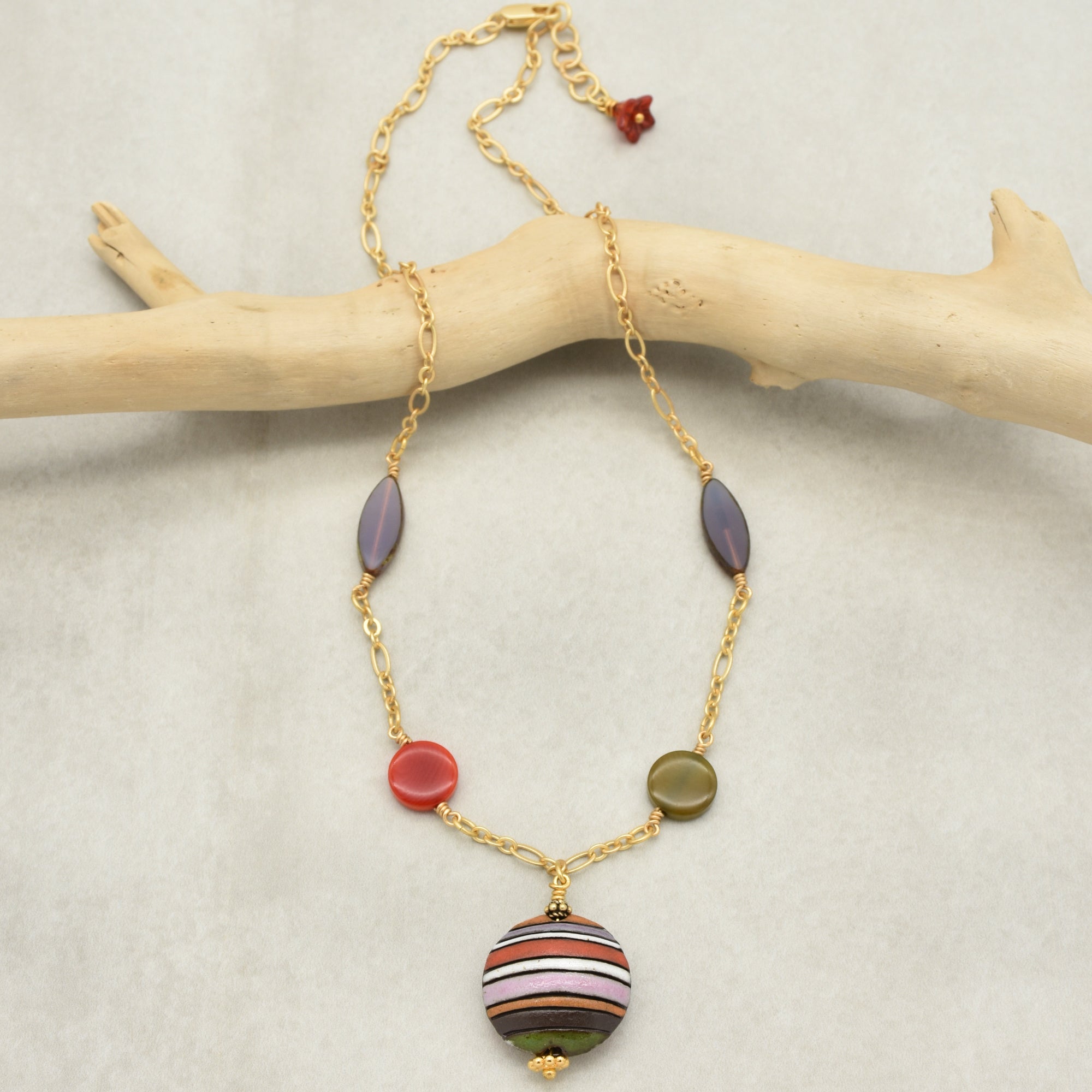 This image shows a necklace. It has a gold chain with colorful beads and a larger circular striped pendant in the center.
