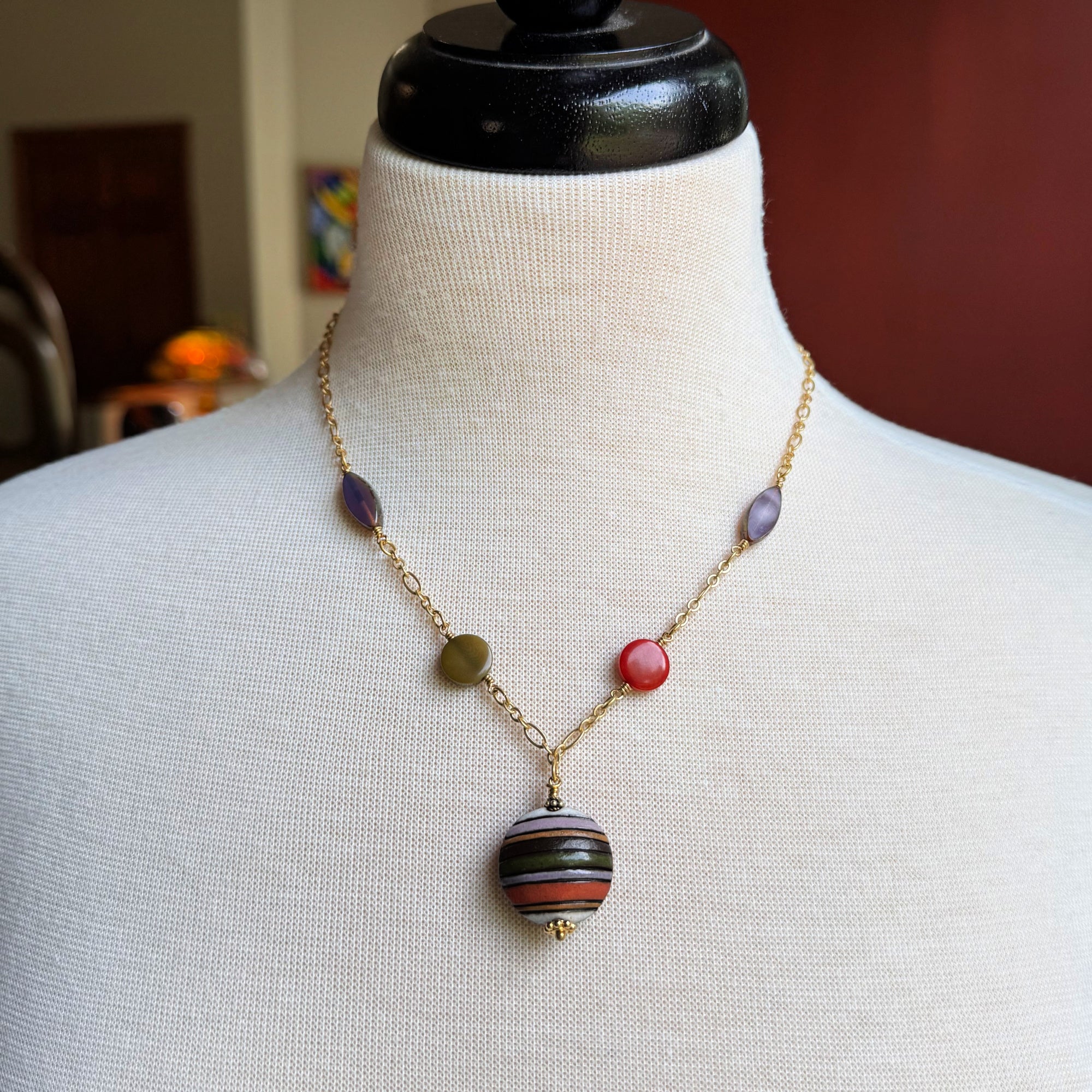 This image shows a necklace. It features a gold chain with colorful beads spaced along it, and a larger, round, striped pendant hanging at the center.