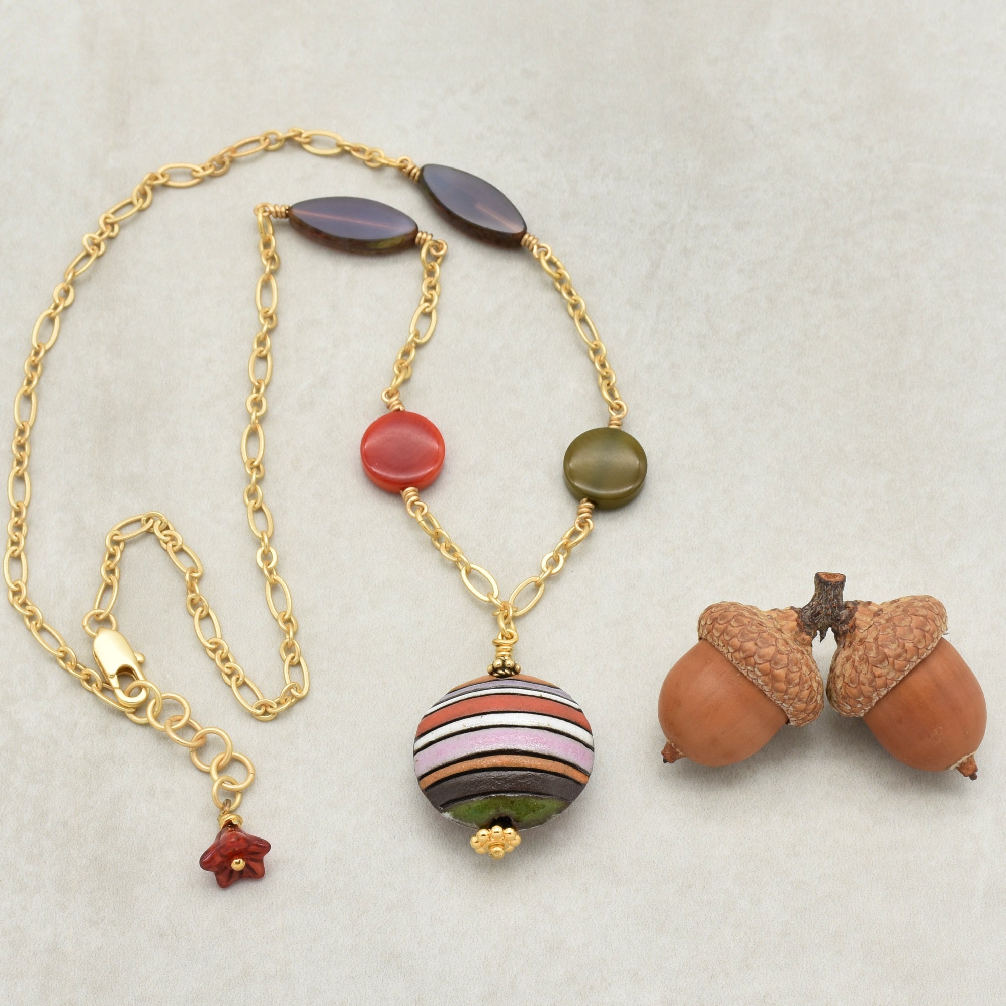 This image shows a necklace. The jewelry piece has a chain with variously shaped and colored beads and a clasp typically found on necklaces. The two acorns to the side are not part of the jewelry.