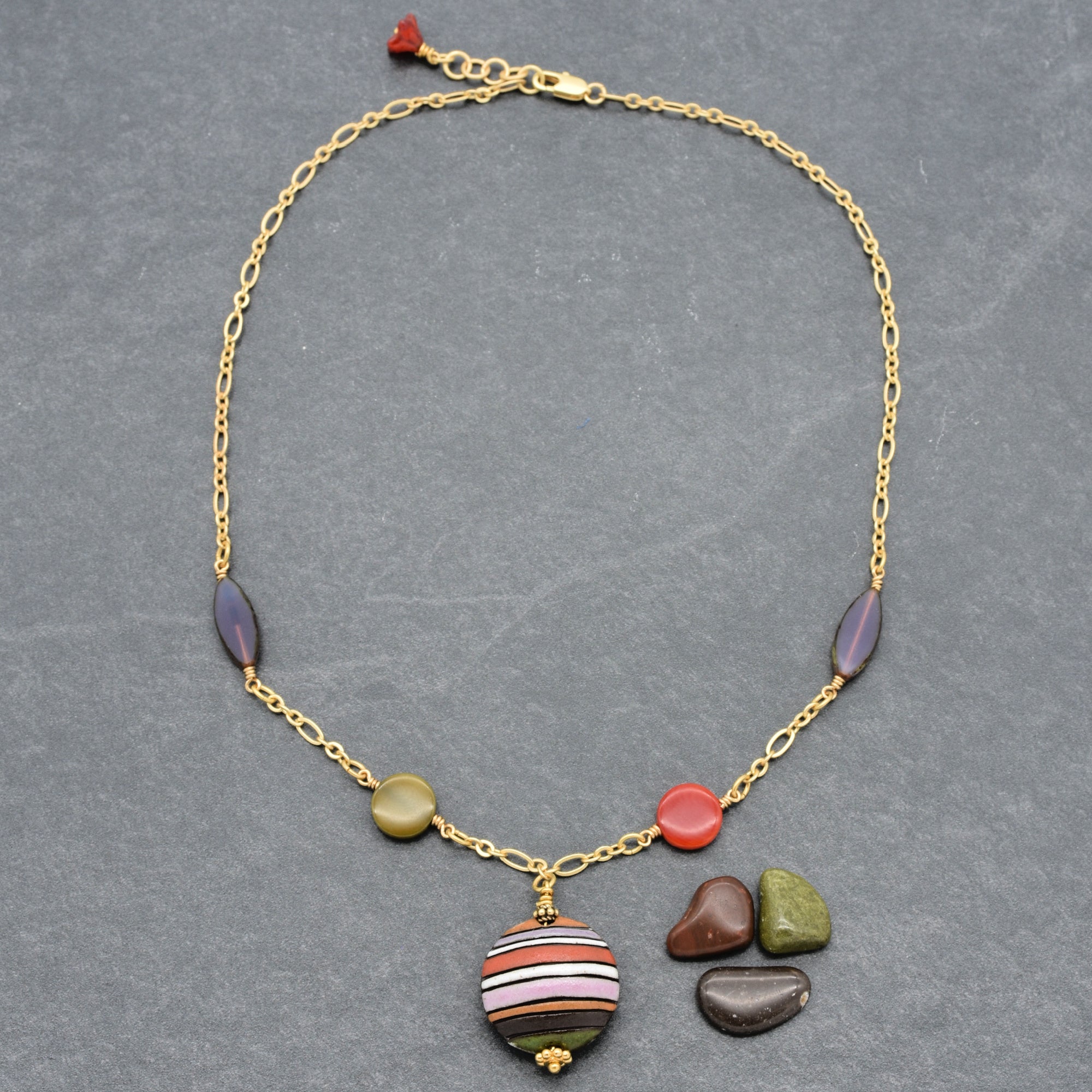This image shows a necklace. It has a gold chain with various beads and stones attached, including a central round striped pendant and other small, differently shaped stones. The length and shape of the jewelry suggest it's designed to be worn around the neck.