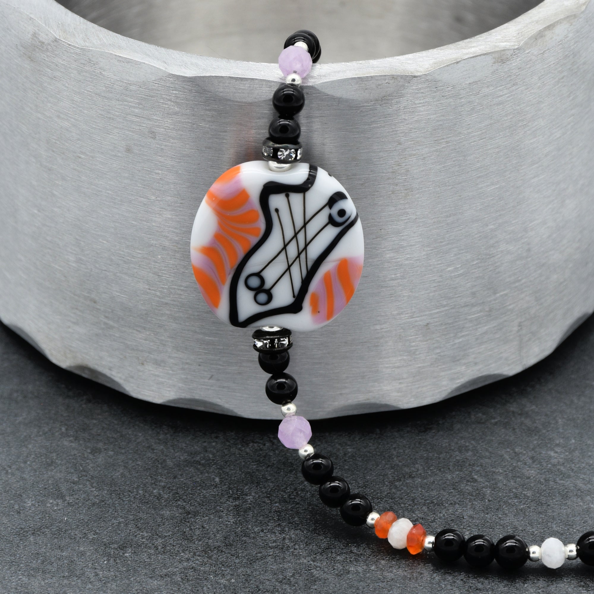 The image shows a piece of jewelry that is a bracelet. It features a round decorative bead with orange, white, and black patterns, surrounded by smaller black beads and some purple and silver accents along the string.