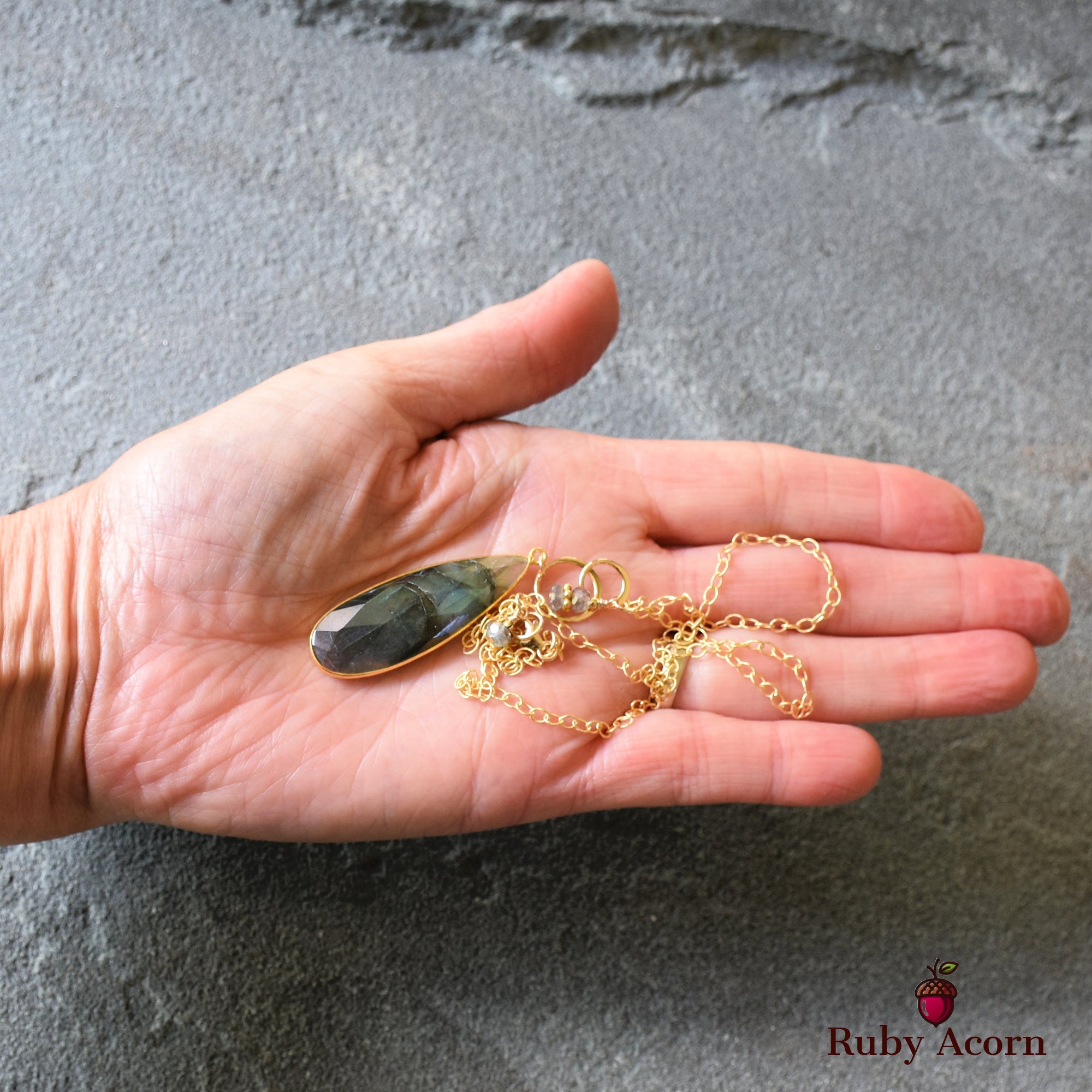 The image shows a necklace. It features a gold chain with a large, elongated teardrop-shaped pendant that appears to be a faceted stone. The necklace is being held in an open palm against a gray textured background.