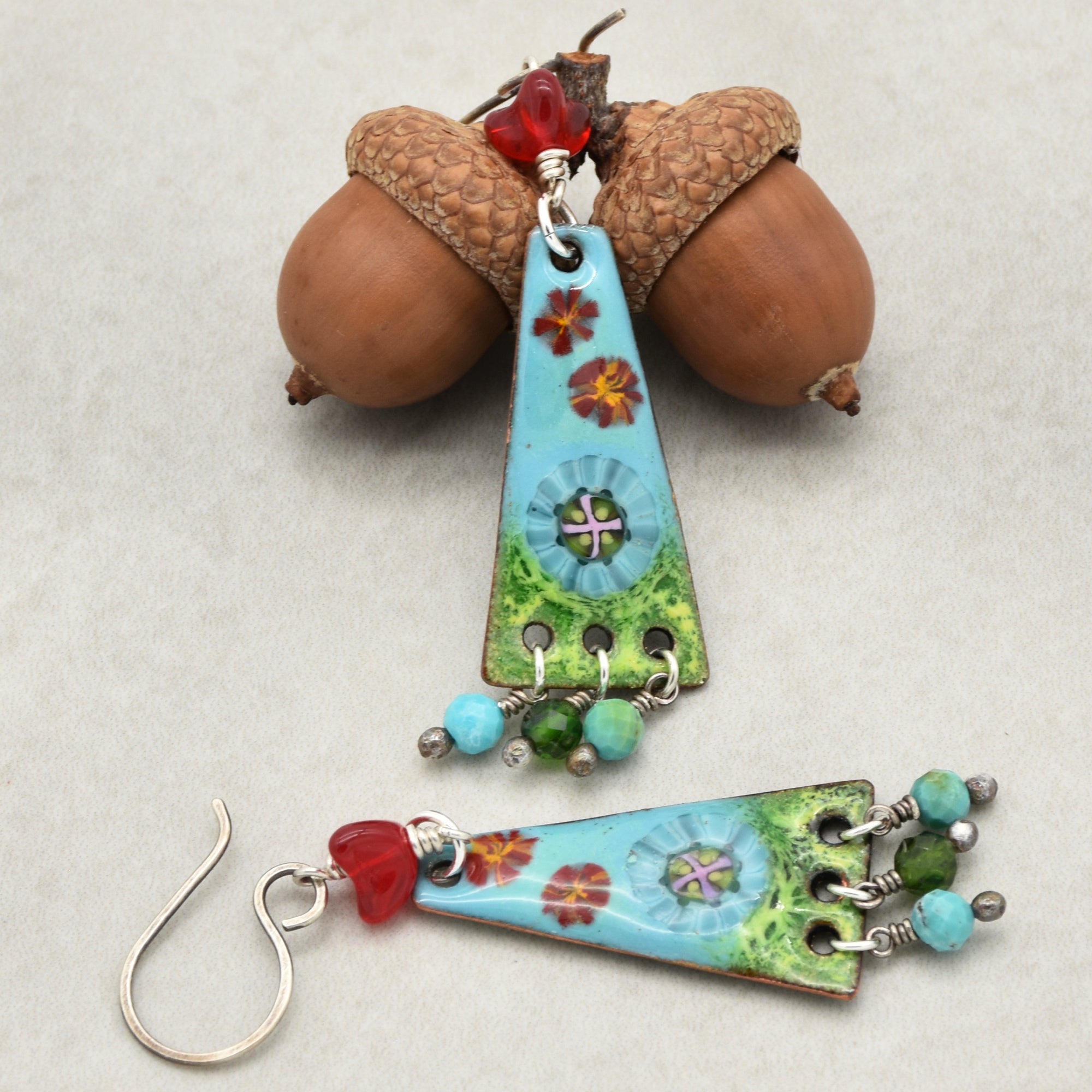 The image shows a pair of earrings. They have a hook for piercing, with decorative triangular pendants featuring colorful designs and small bead dangles at the bottom.