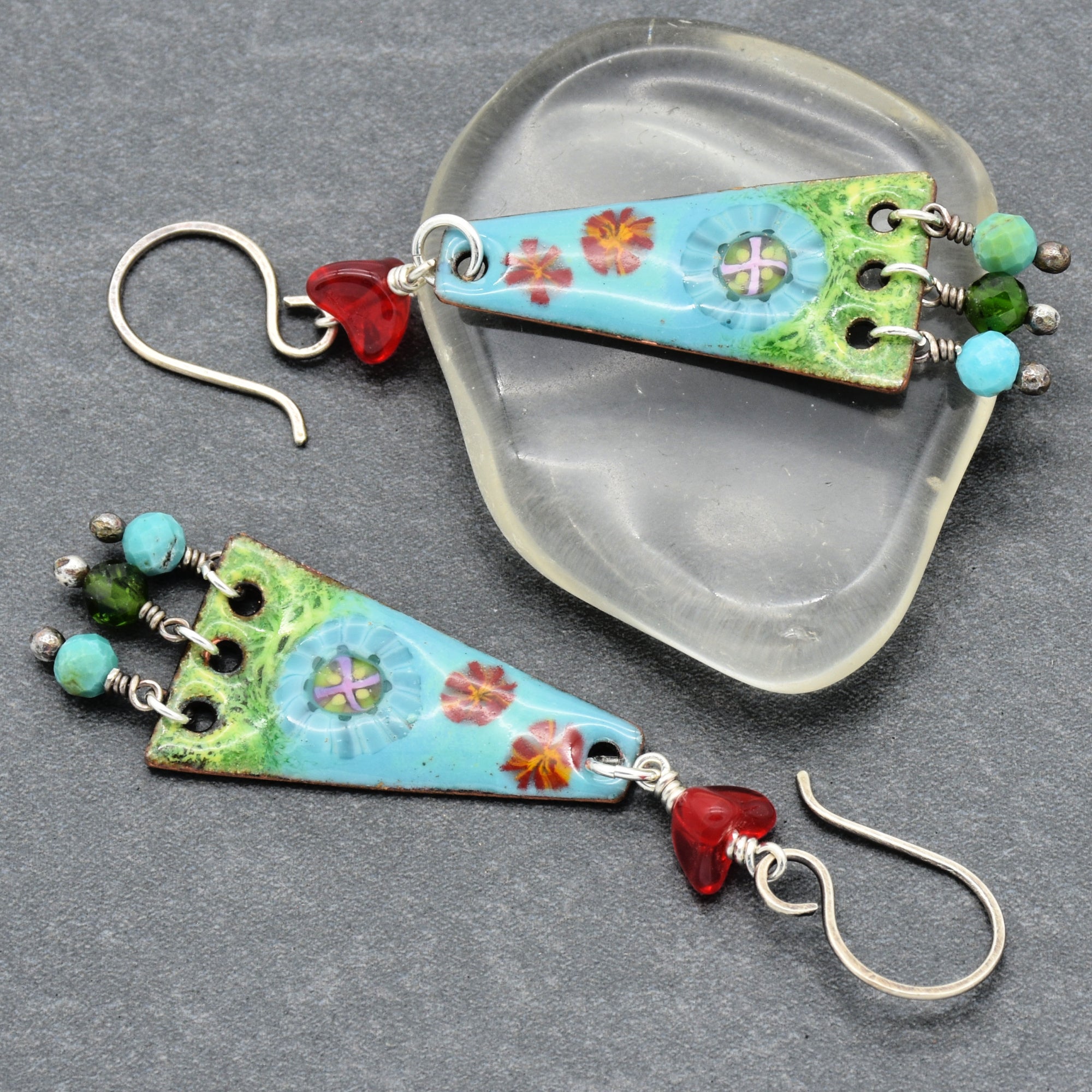 The image shows a pair of earrings. The jewelry has a hook design for the ear, with colorful, decorative triangular pendants featuring small beads and floral patterns.