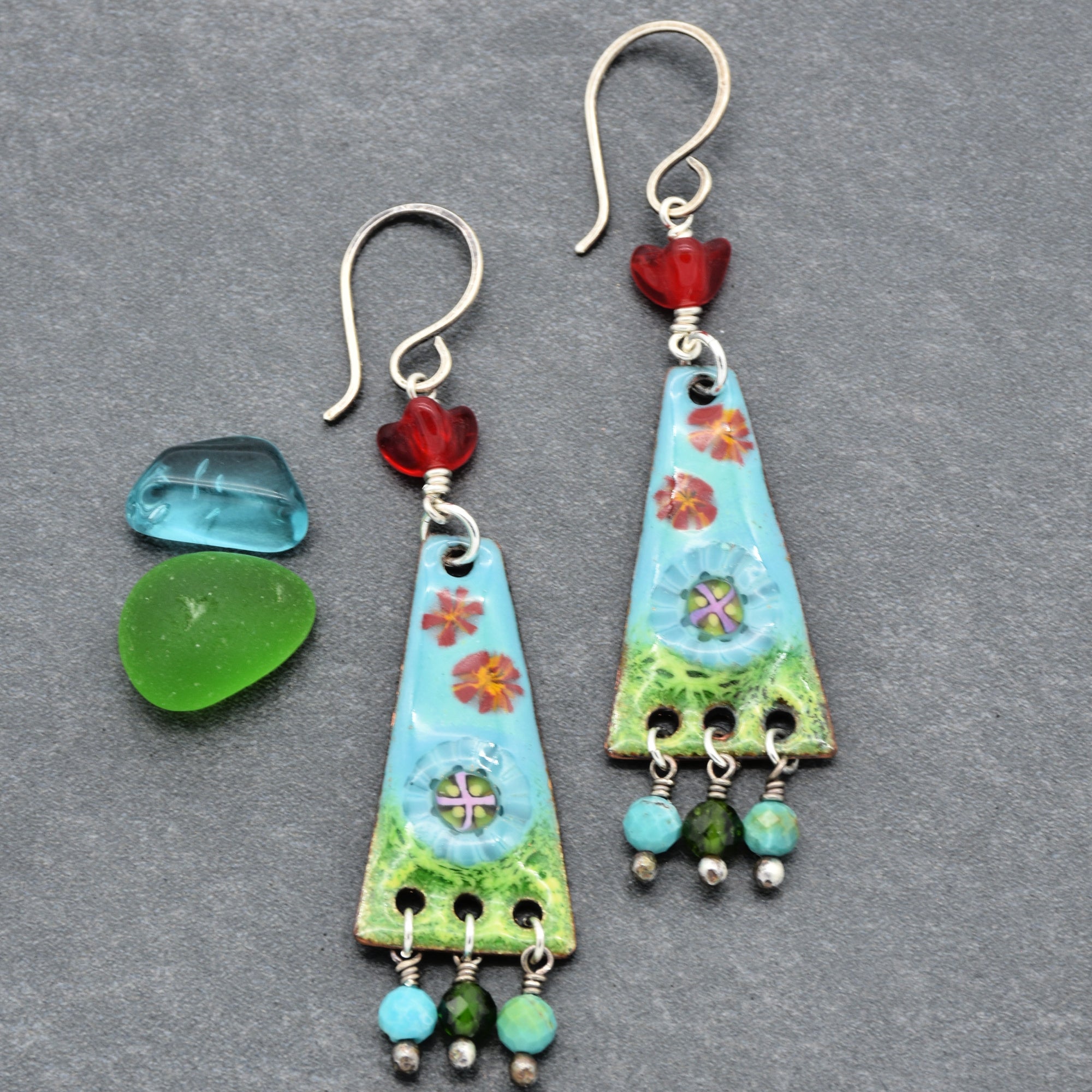 This image shows a pair of earrings. They have hooks for placing through pierced ears and decorative elements hanging from them including red, blue, and green beads with a floral design on a triangular base.
