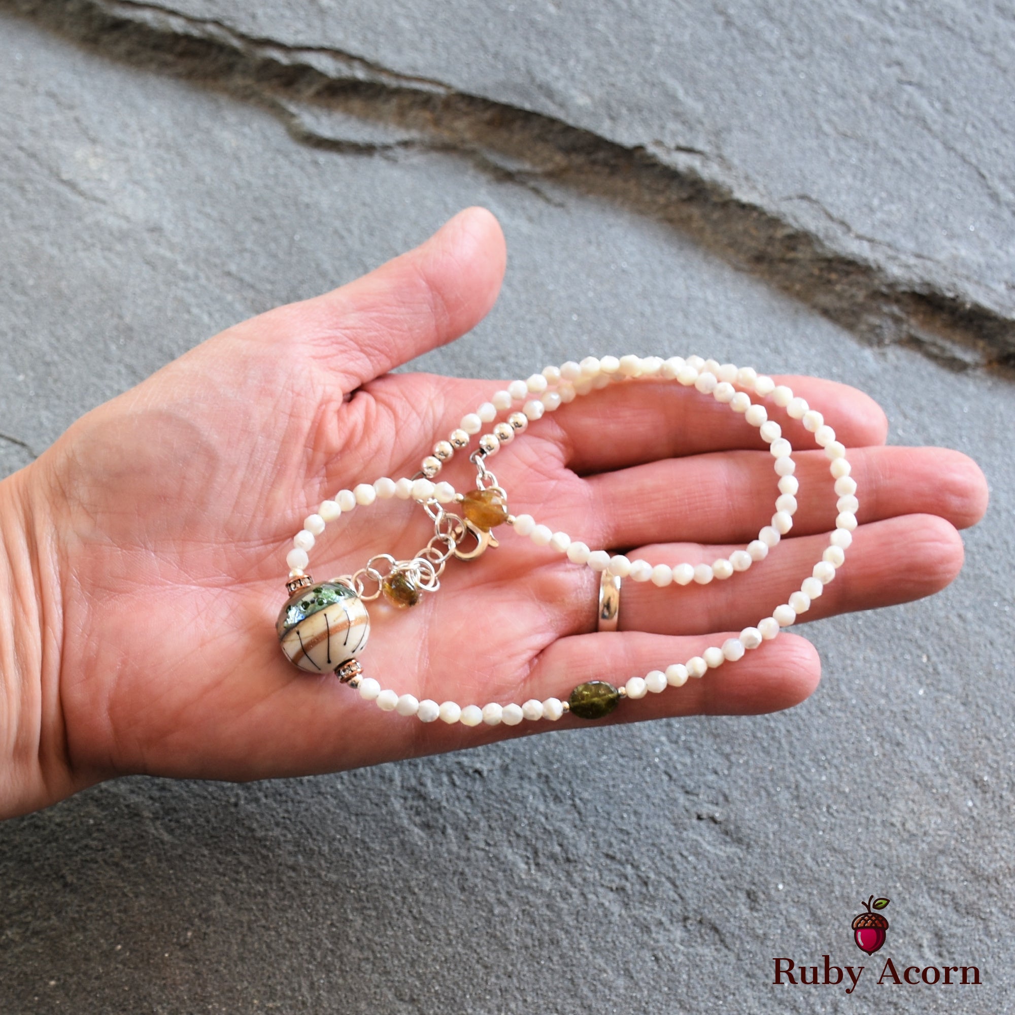 This image shows a bracelet. The jewelry piece is a beaded design with small white beads and a larger decorative bead, along with some additional smaller accent beads. It is being held in a person's hand. The logo in the corner says "Ruby Acorn."