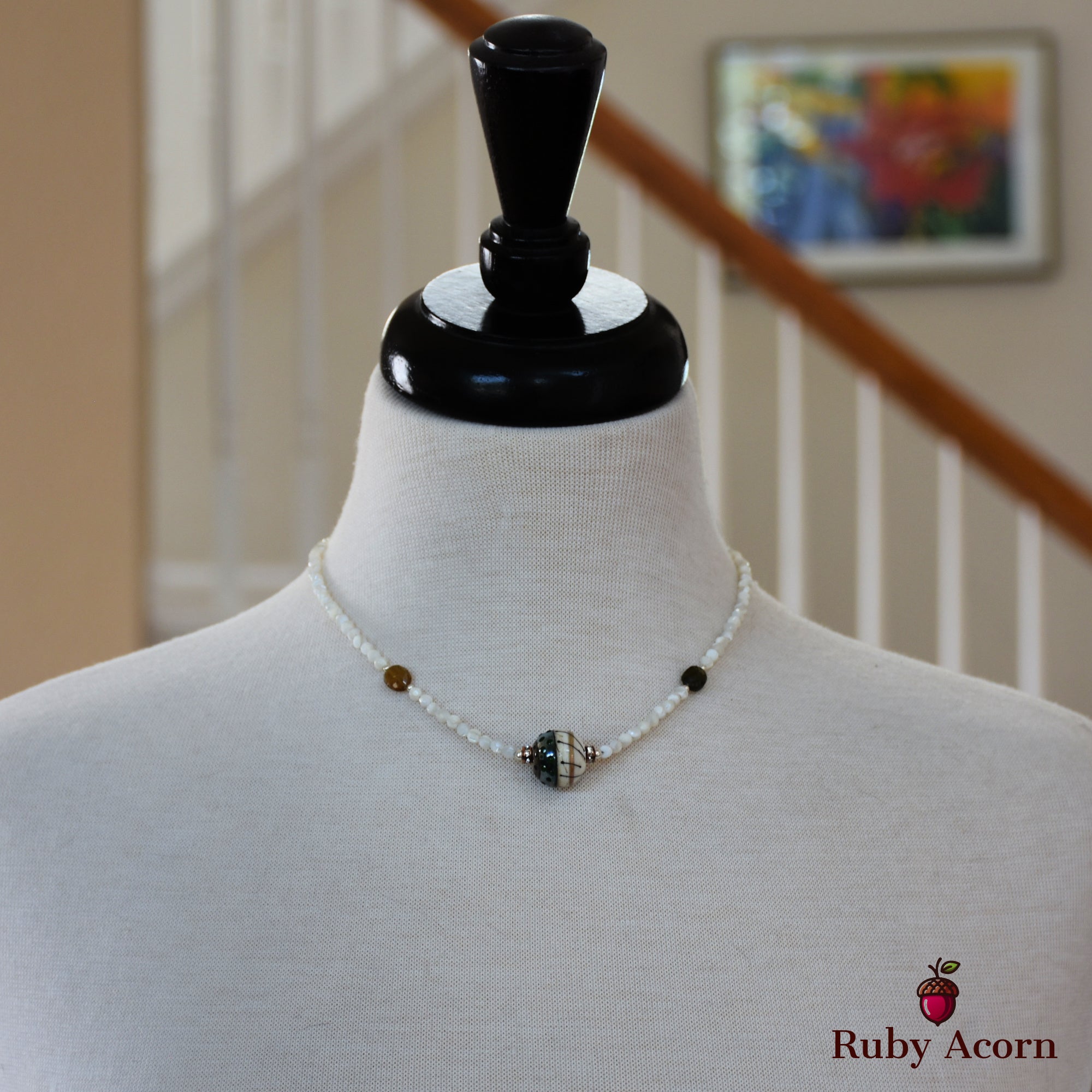 This image shows a necklace. It is displayed on a mannequin bust and consists of small white beads with a larger, more decorative bead in the center.