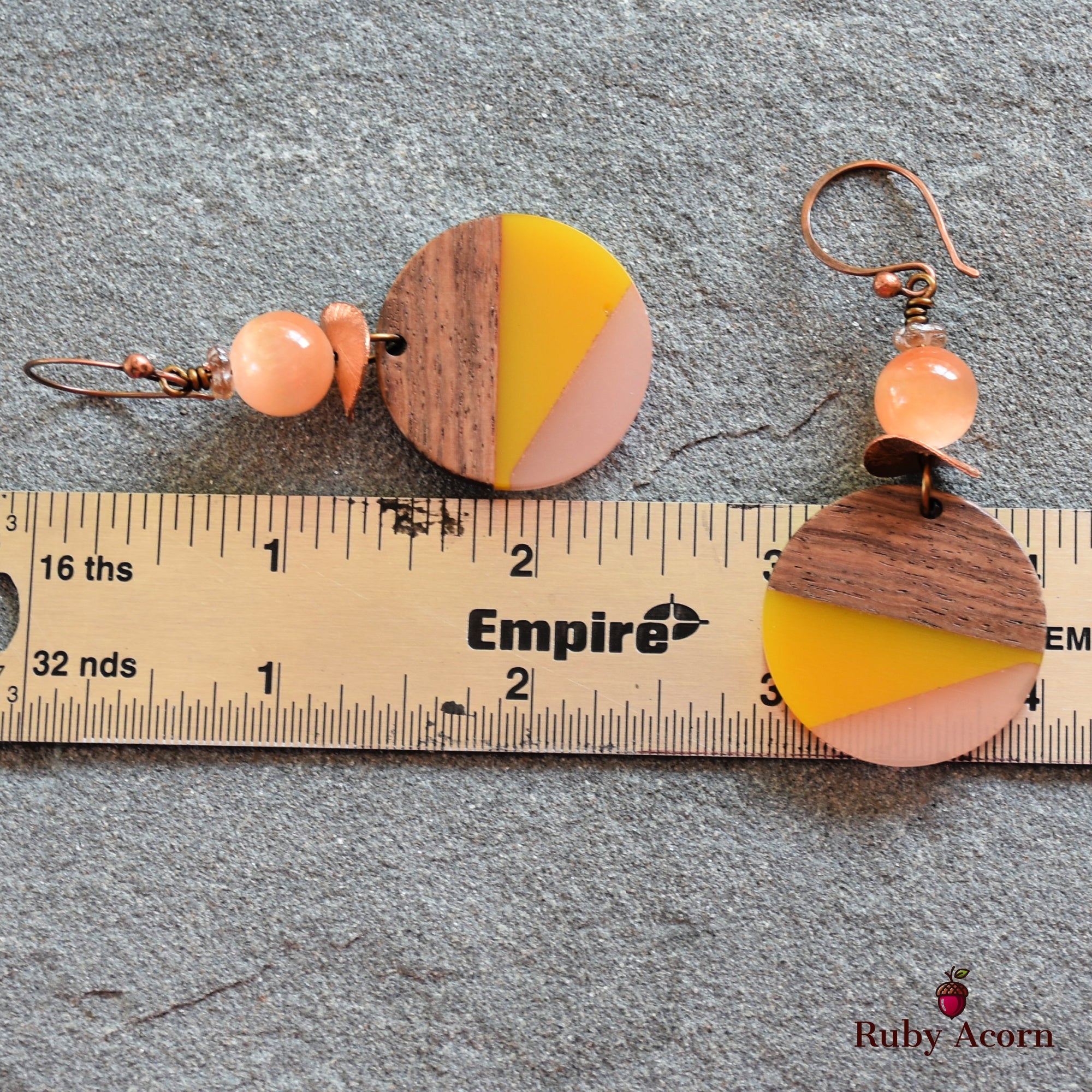 The image shows a pair of earrings. They have hooks for wearing through pierced ears and feature circular decorative pieces made of wood and colored sections in yellow and pink, along with a peach-colored bead above the circular part. The ruler in the image indicates their size.