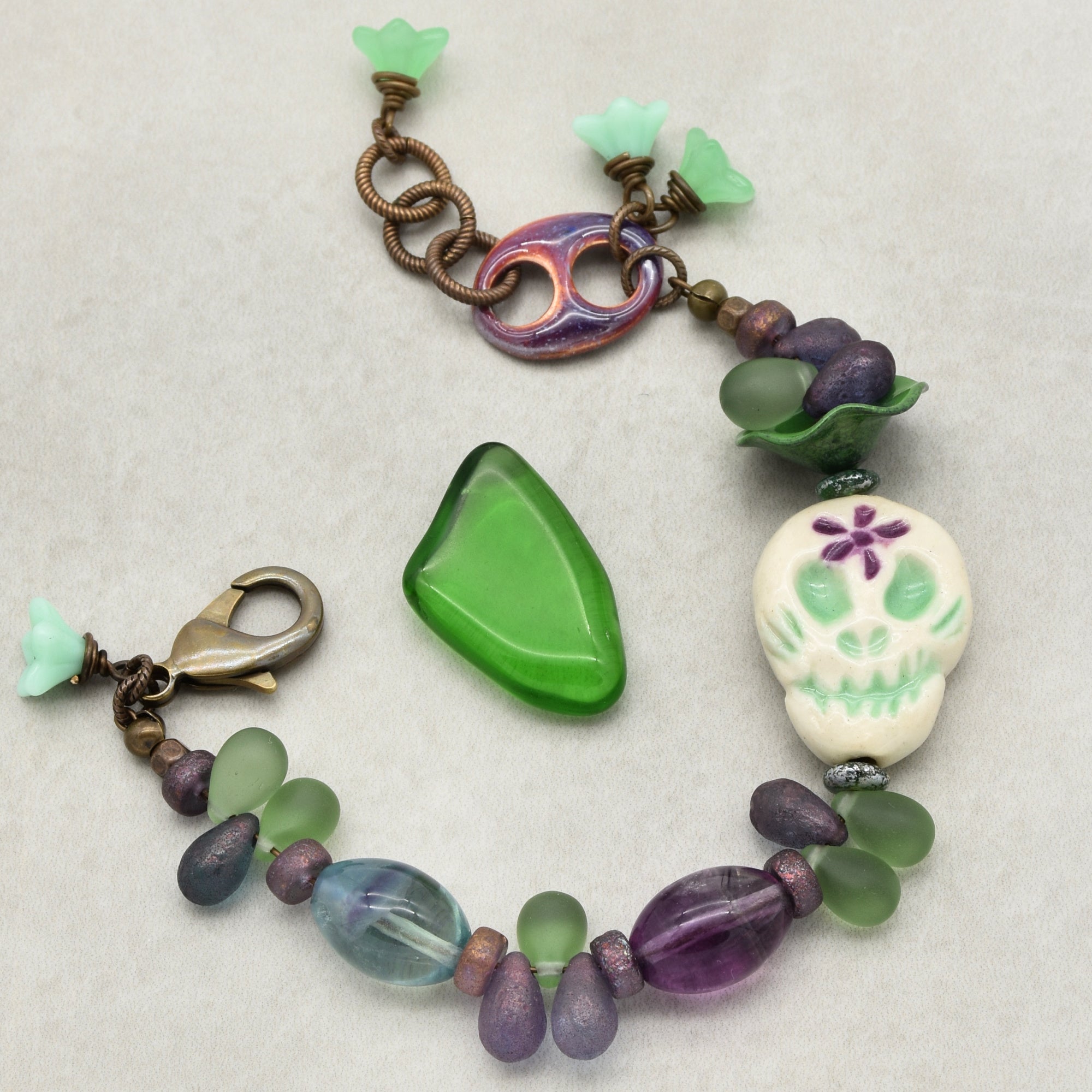 The image shows a bracelet. It has beads of different shapes and colors, including green, purple, and some with a metallic finish. One of the prominent features on the bracelet is a white skull bead with green and purple detailing. The bracelet also has a lobster clasp for fastening.