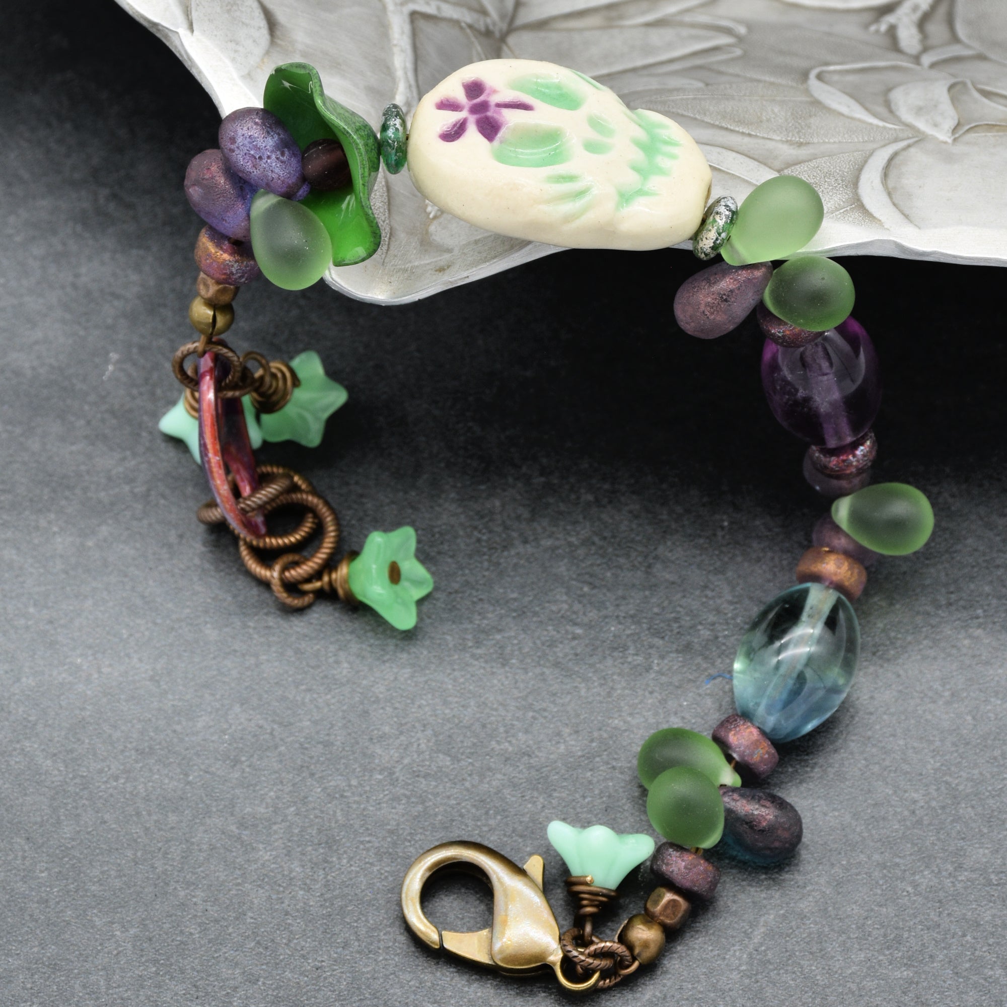 This image shows a bracelet. It features various colorful beads, including green frosted beads, purple and blue beads, and a central bead shaped like a skull with green and purple designs. The bracelet also has a clasp for fastening.