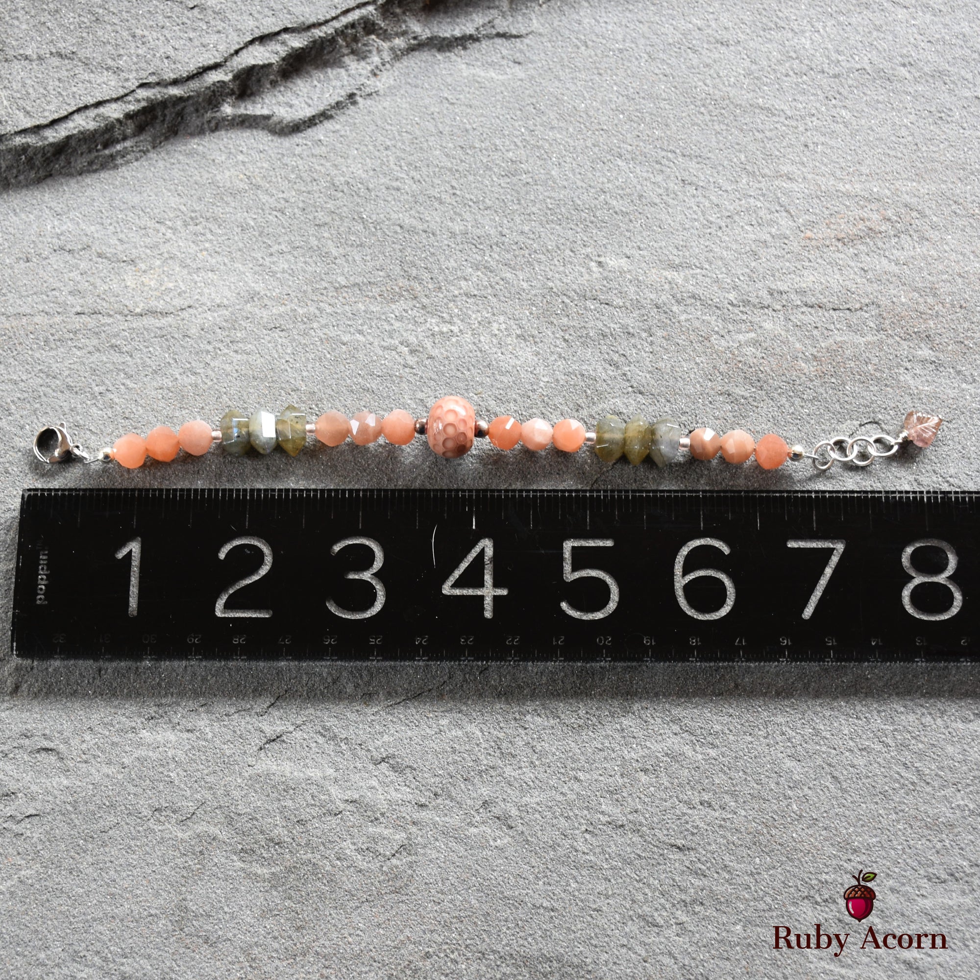 This image shows a bracelet. It is relatively short in length, approximately 7 to 8 inches, which is typical for a bracelet. The jewelry has various beads, including round peach-colored beads and some irregularly shaped gray-green beads, with a clasp on one end and an adjustable chain on the other. The ruler placed beneath it helps indicate the size.