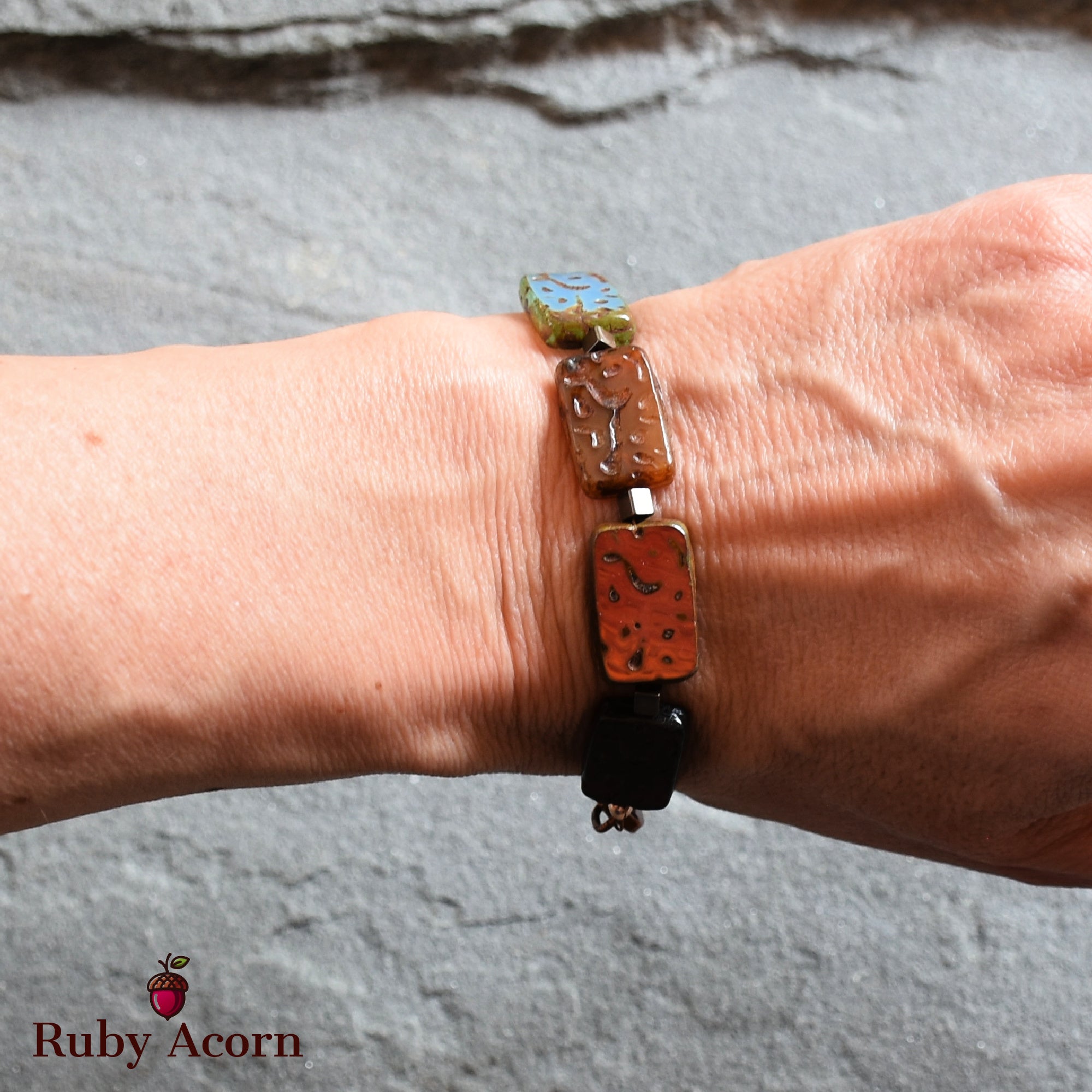 This image shows a bracelet. It consists of rectangular beads in various earth tones threaded together and worn around a wrist.