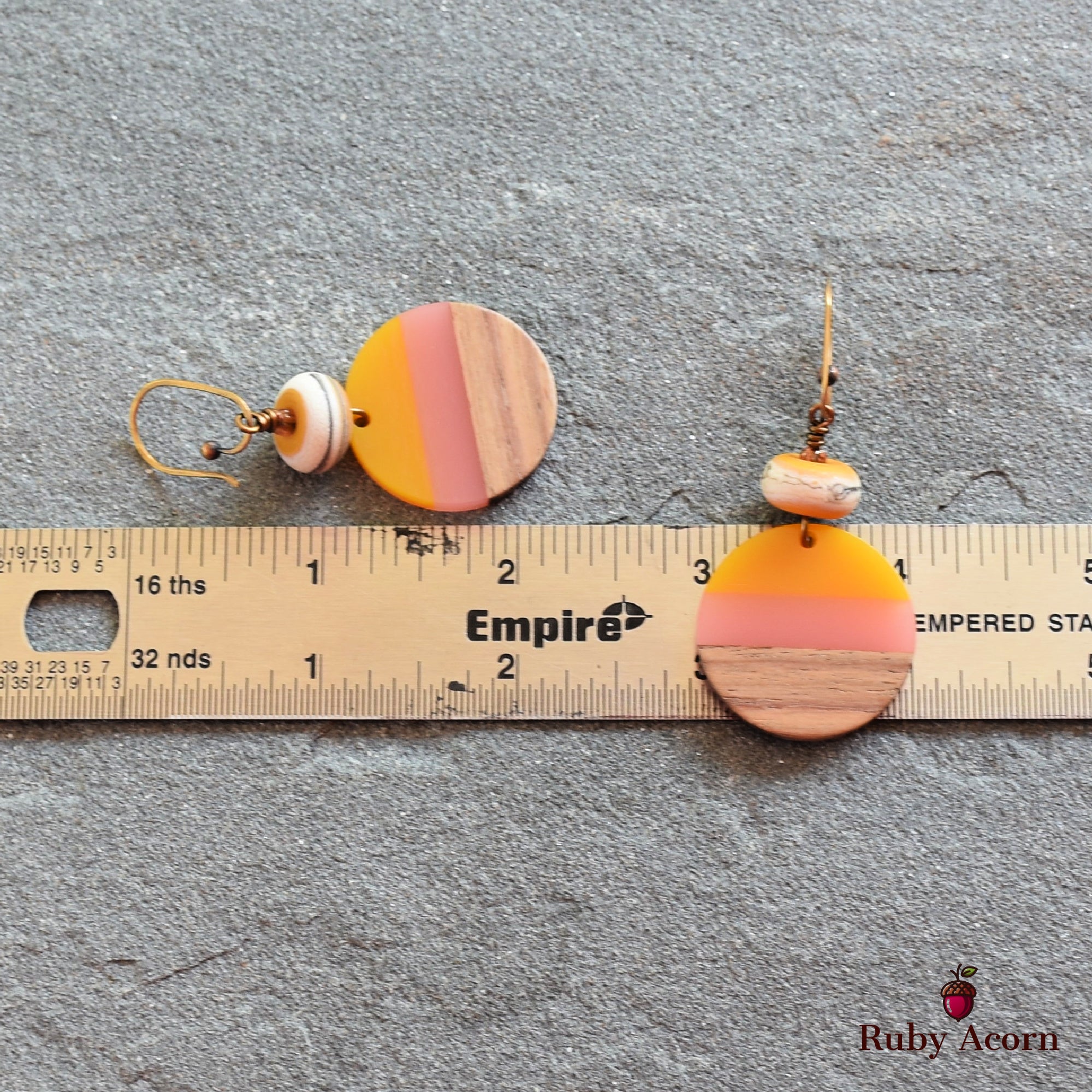 The image shows a pair of earrings. They are circular with a striped design in yellow, pink, and wood-tone colors, and have a hook for wearing. The ruler beneath them indicates their size.
