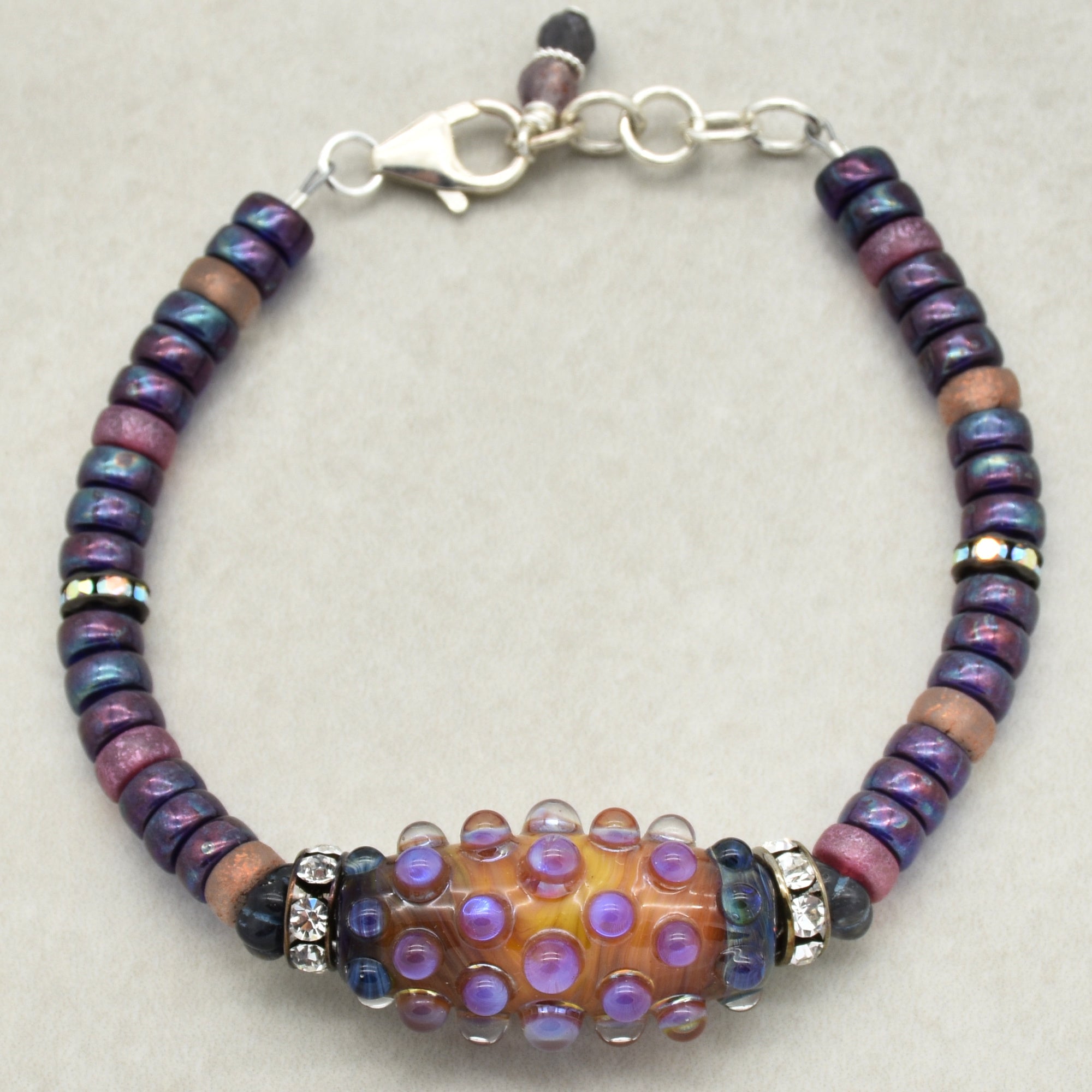 This image shows a bracelet. It features a central decorative bead with raised purple dots, surrounded by smaller beads in shades of purple, blue, and pink, and it has a clasp for fastening. The size and design are consistent with a bracelet.