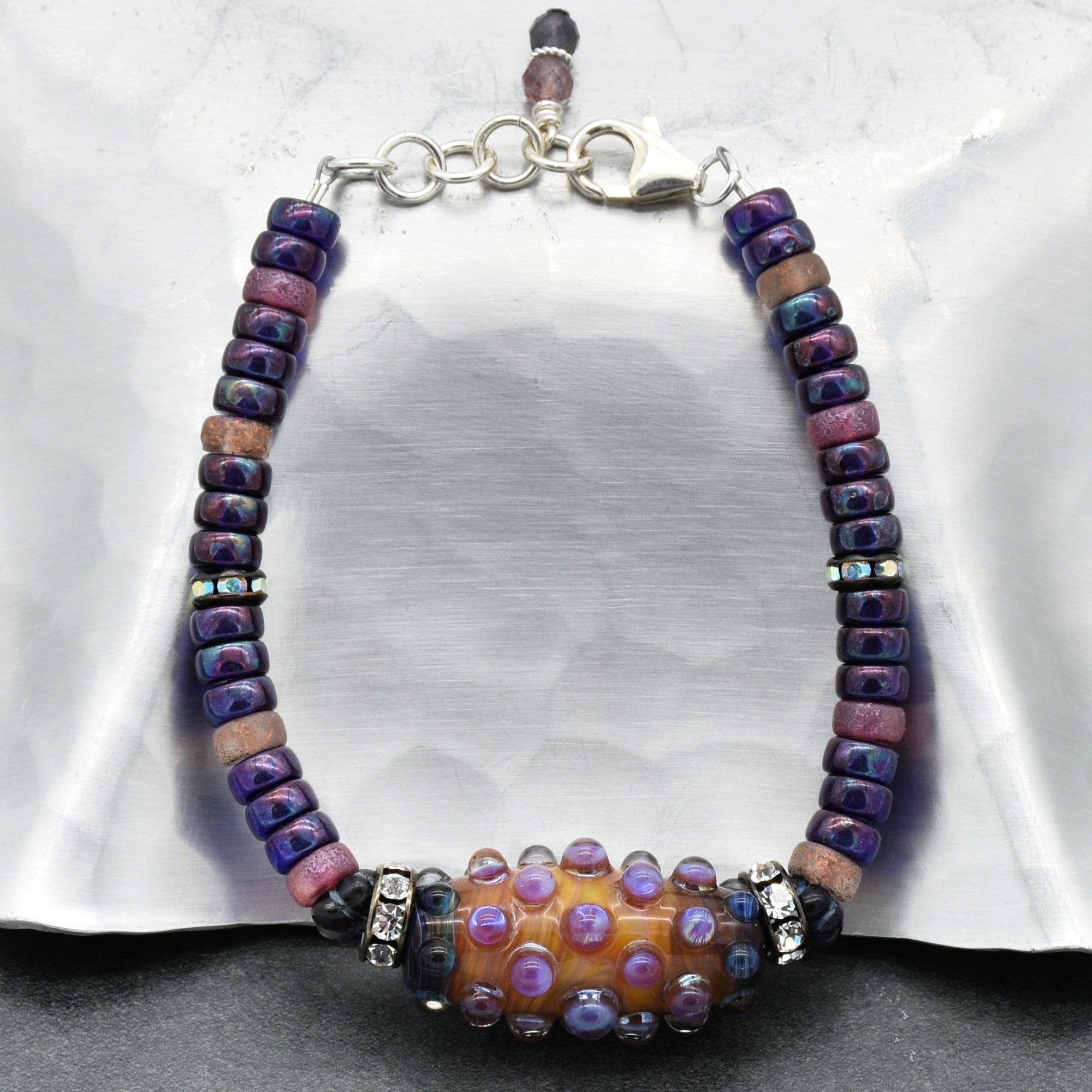 This image shows a bracelet. The jewelry piece is characterized by beads arranged in a circular band with a clasp, which is typical of bracelets. The beads are of various colors including purple, brown, and a central larger bead with decorative raised dots and rhinestone spacers.