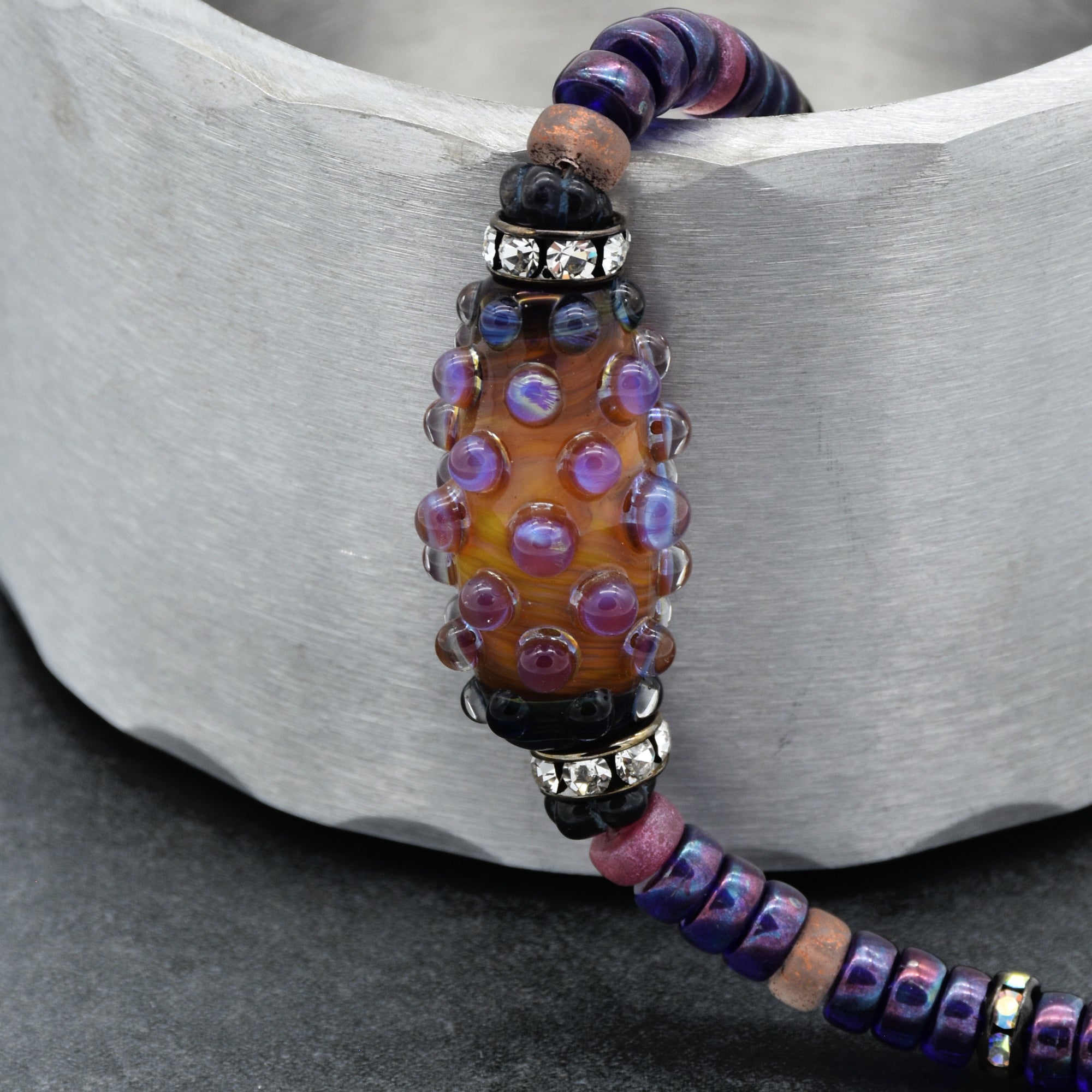 The image shows a necklace featuring a large central bead with raised purple dots, flanked by smaller beads in shades of purple, black, and pink. There are also some clear, sparkly spacer beads near the central bead.