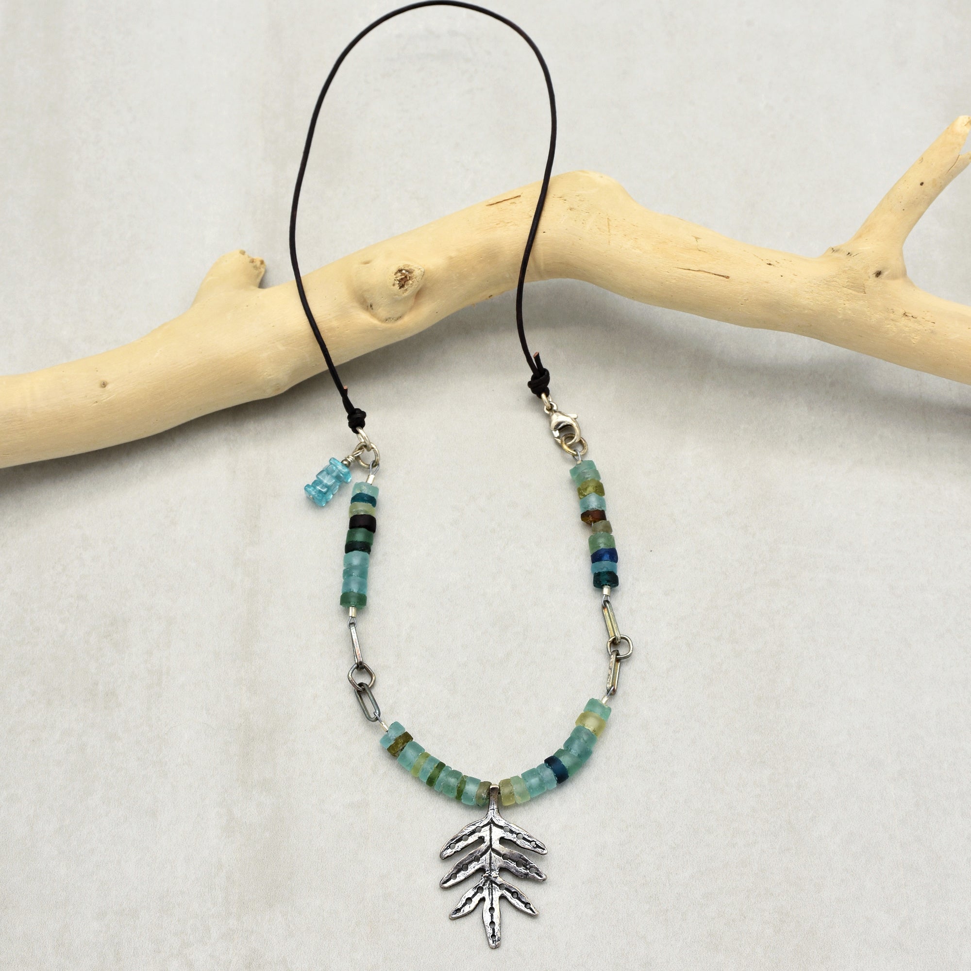 The image shows a necklace. It has a black cord with beads in various shades of blue and green, and a silver pendant in the shape of a leaf or branch hanging in the center.
