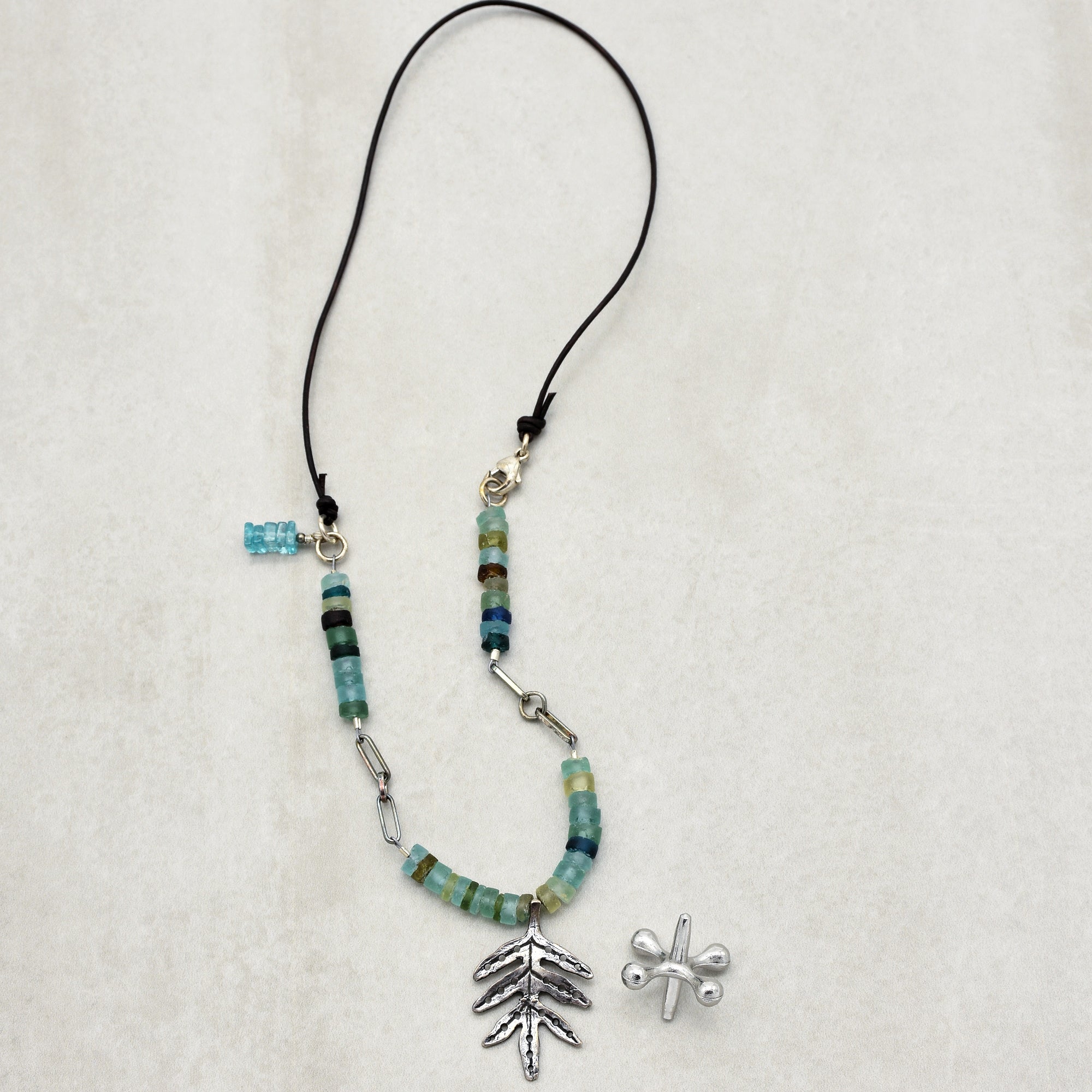 The image shows a necklace. The necklace has blue and green beads and a silver leaf-shaped pendant, strung on a black cord.