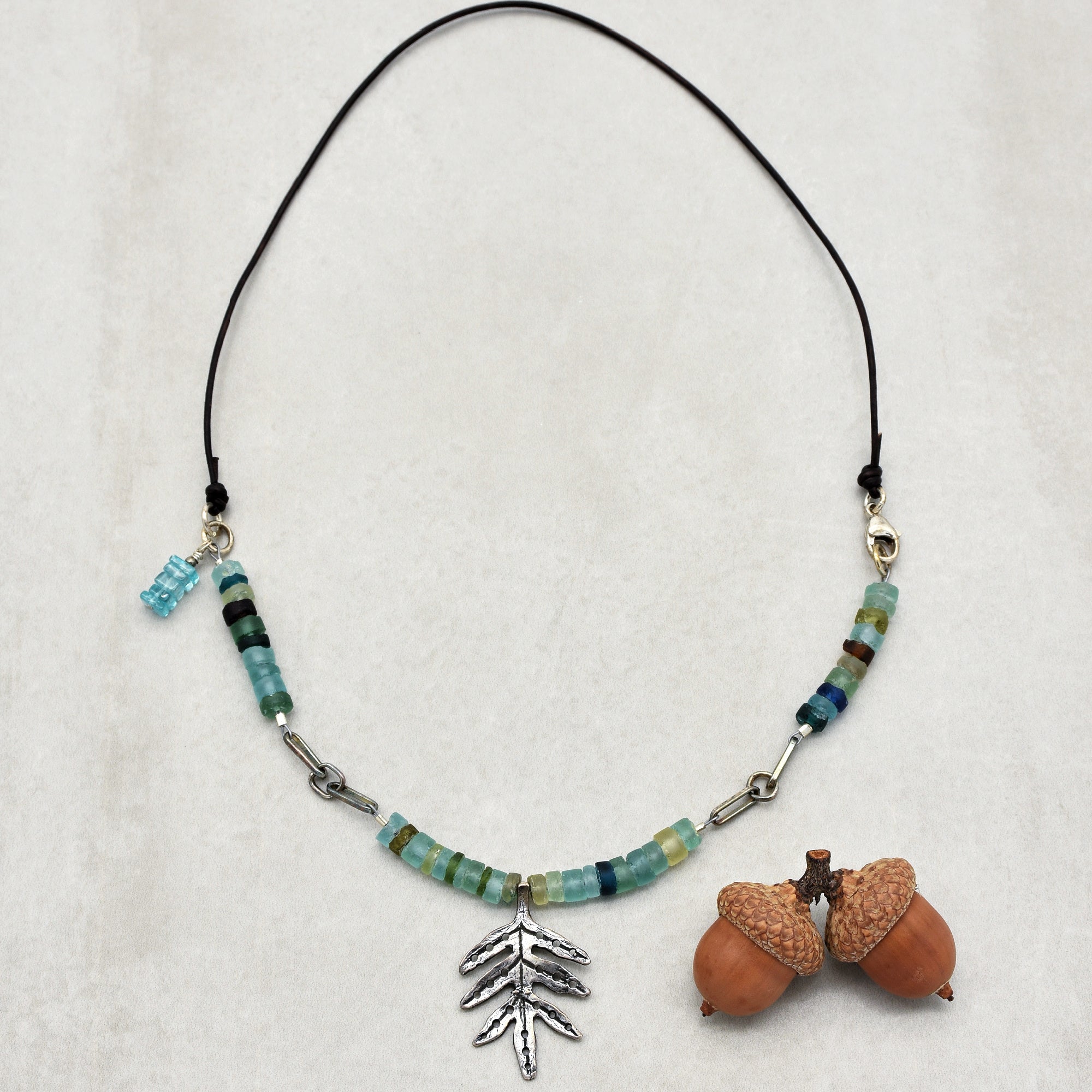This image shows a necklace. The jewelry piece has a string with beads and a metal leaf-shaped pendant hanging in the center. The length and style indicate it is meant to be worn around the neck.
