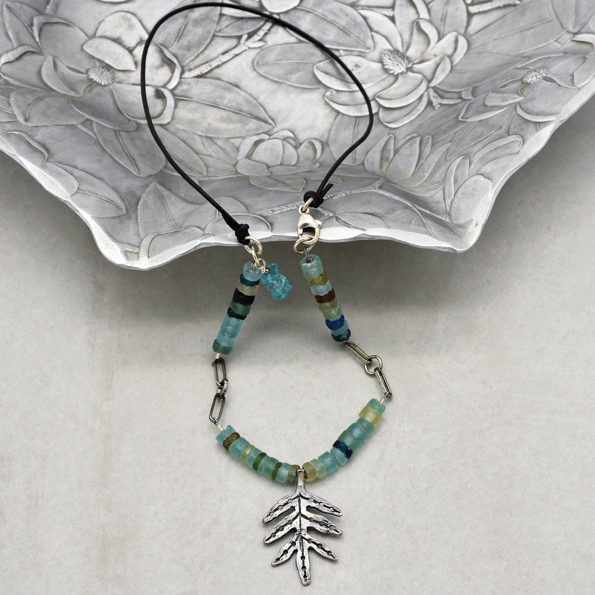 This image shows a necklace. It has a circular band made of a dark cord with a clasp, and it features beads and a decorative leaf-shaped pendant hanging from it.