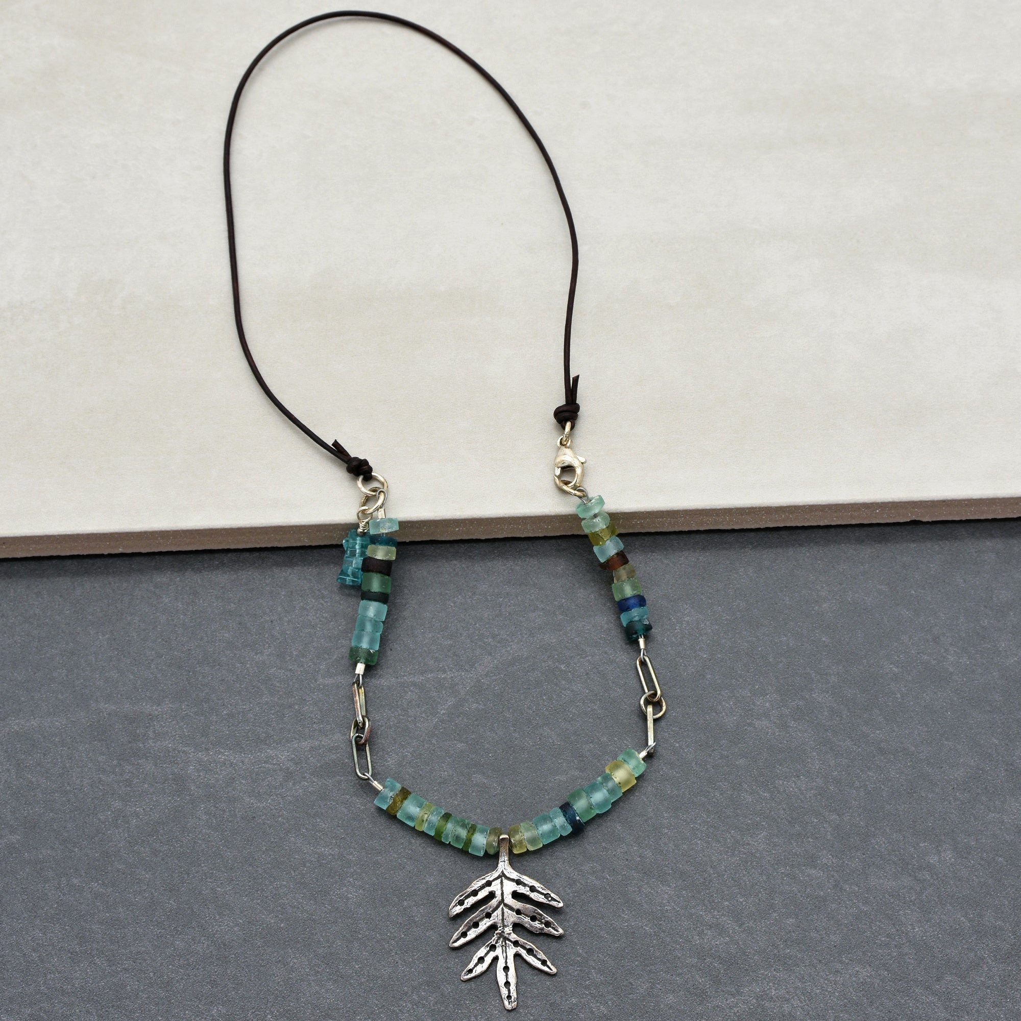 This image shows a necklace. It has a string with beads and a leaf-shaped pendant hanging from it. The clasp at the top further indicates that it is meant to be worn around the neck.