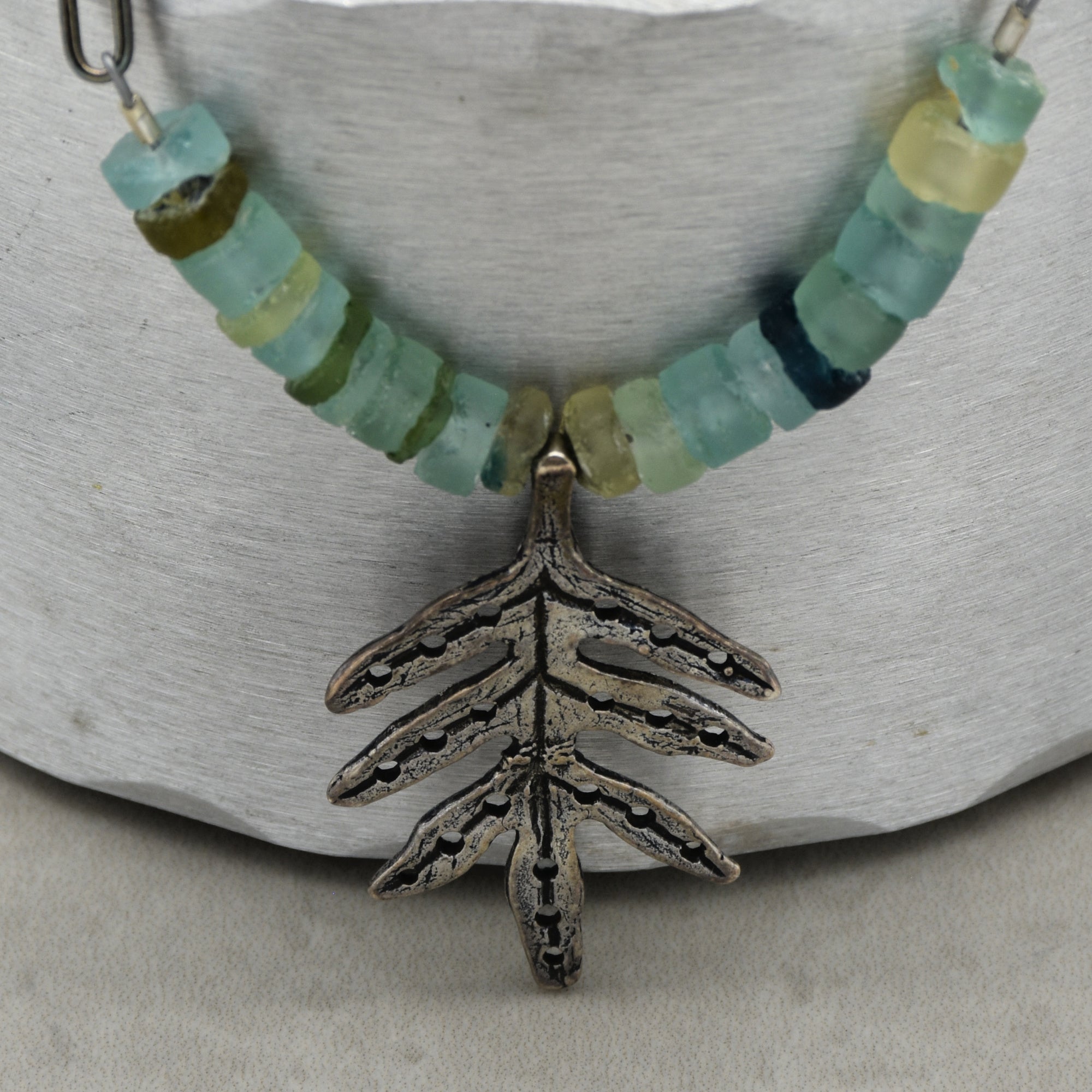 This image shows a necklace. It has a series of light blue and green beads strung together and features a pendant shaped like a leaf or a plant branch.