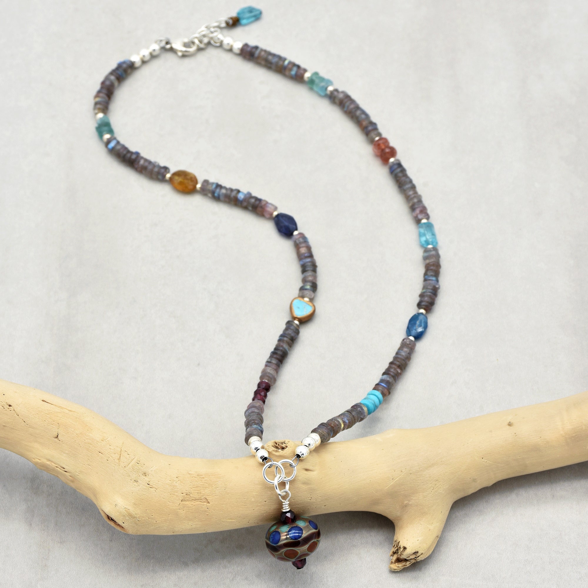 The image shows a necklace. It features a string of beads in various colors and shapes, with a pendant hanging at the center. The necklace is arranged on a light background, with part of it draped over a piece of driftwood or a similar natural object.