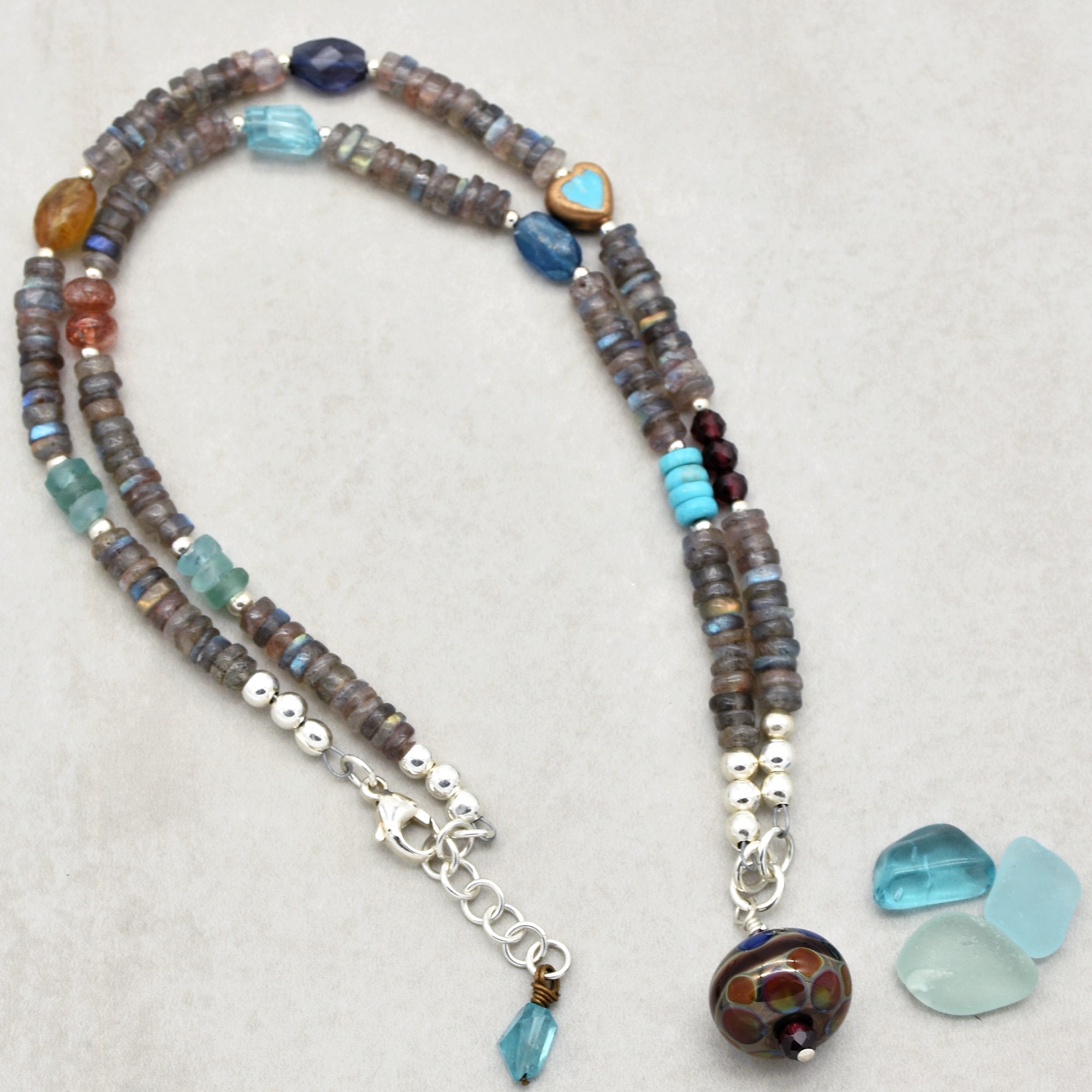 This image shows a necklace. It has two strands of beads with various colors and shapes, including some round silver beads and a larger decorative bead at the end. There is also a clasp for fastening, which is typical for a necklace.