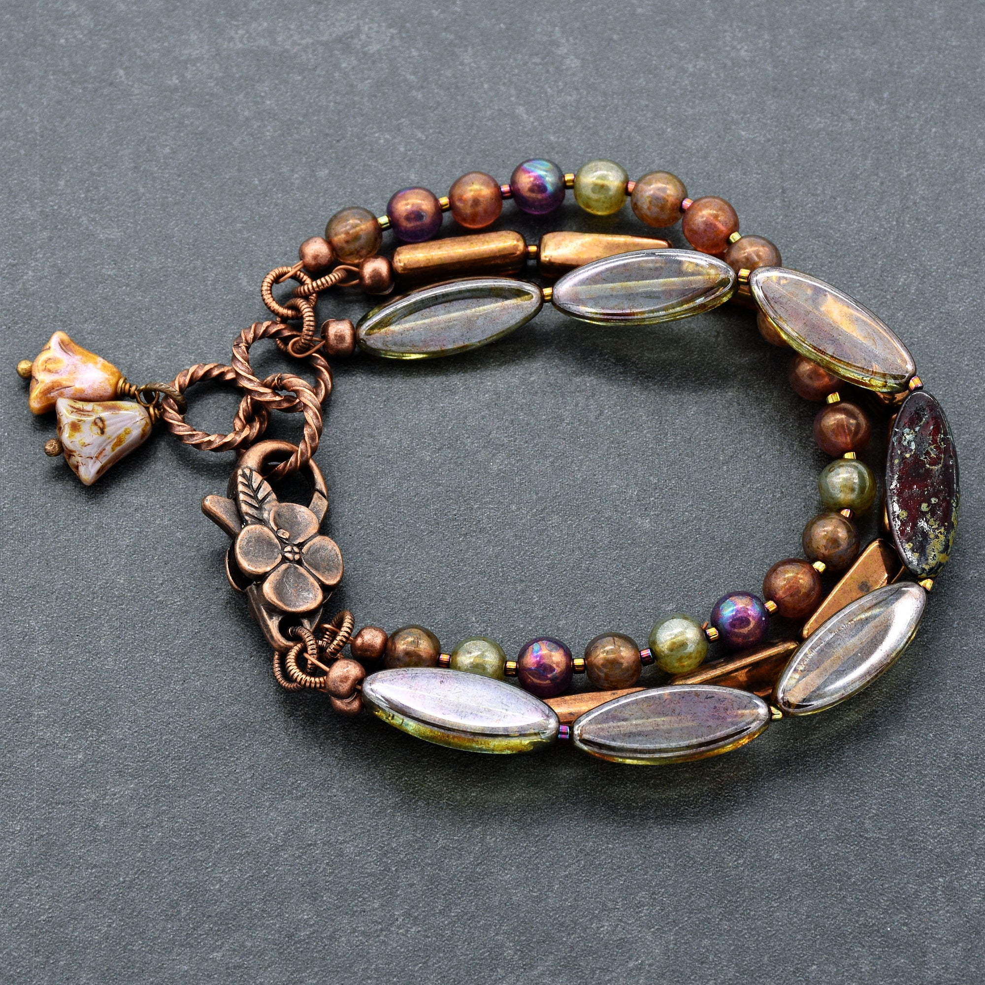 This image shows a bracelet. It features multiple strands with a mix of beads, including round, cylindrical, and oval shapes, and a clasp with a floral design. The bracelet has earthy tones with some iridescent beads.