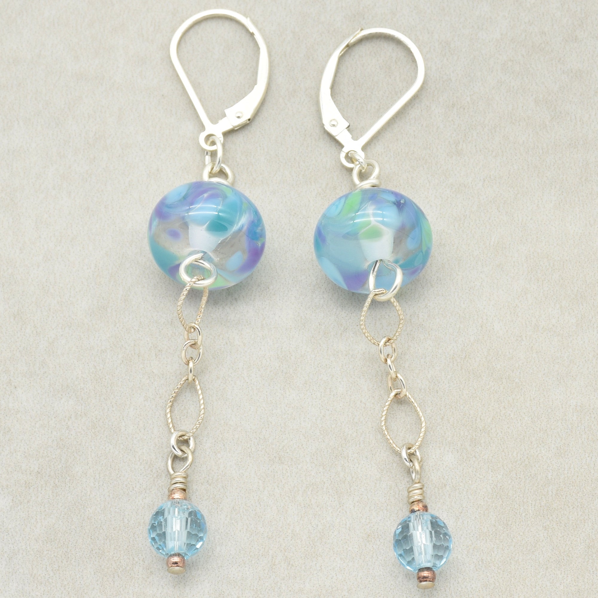 This image shows a pair of earrings. They feature a colorful round bead near the top and a smaller blue bead hanging at the bottom, connected by delicate chain links. The hook style closure confirms that they are earrings.