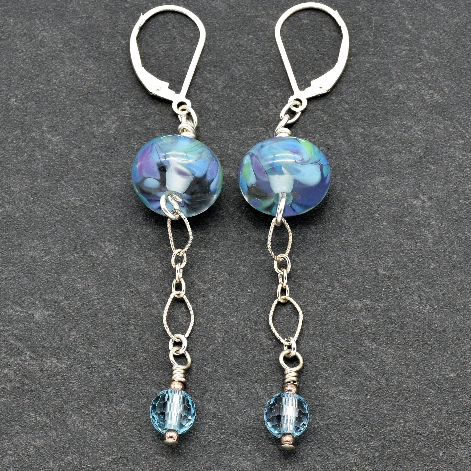 The image shows a pair of earrings. They feature a design with a colorful round bead near the top, connected by a chain of small interlinked loops, and ending with a smaller faceted blue bead at the bottom.