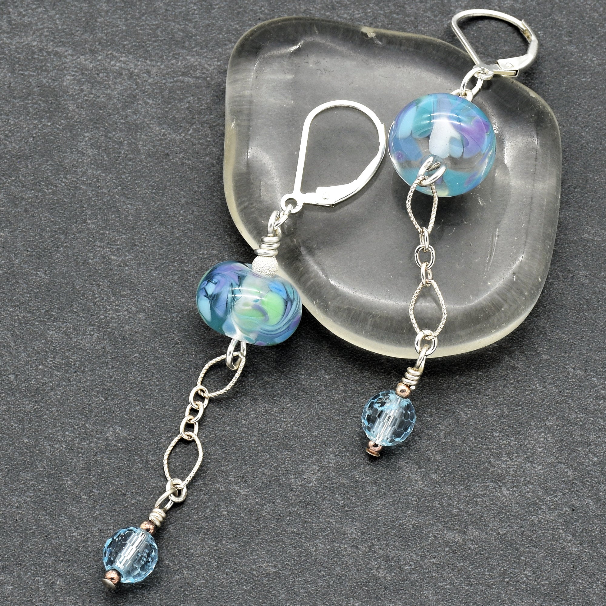 This image shows a pair of earrings. You can tell because of the ear hooks at the top, which are designed to be worn through pierced ears. The design includes blue and purple beads with dangling elements.