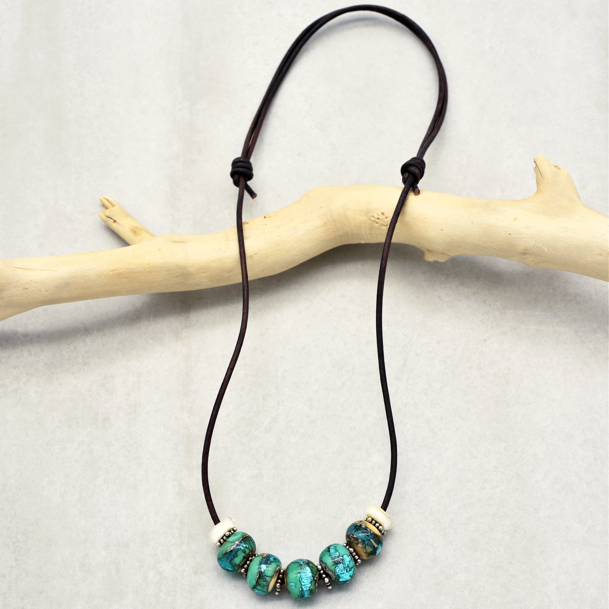 This image shows a necklace. It features a leather cord with turquoise-colored and metallic beads strung along the center. The cord has knots on either side of the beads to keep them in place.