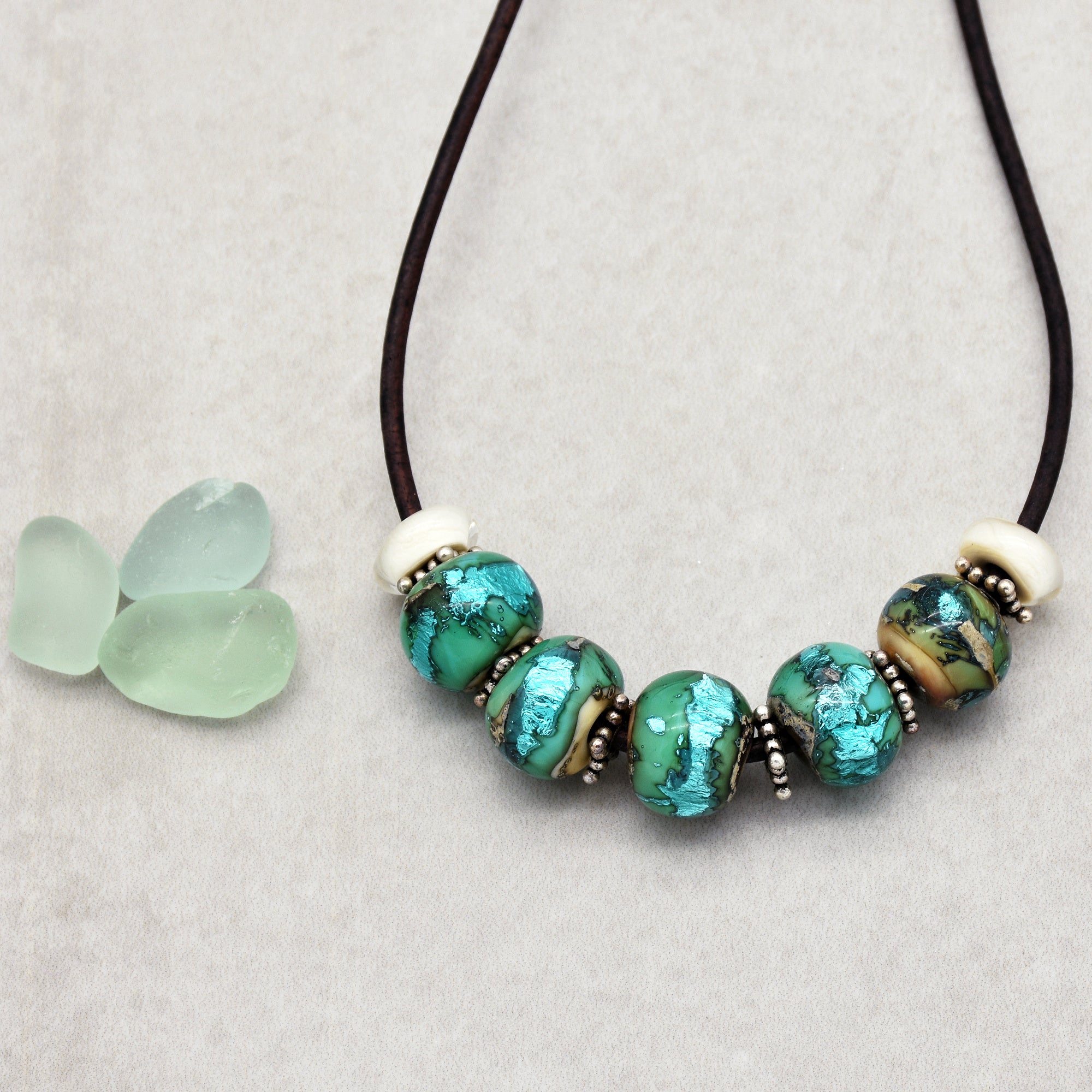 This image shows a necklace. It has a brown cord and features several large, round beads with a mix of green, blue, and metallic colors, interspersed with small silver spacer beads.